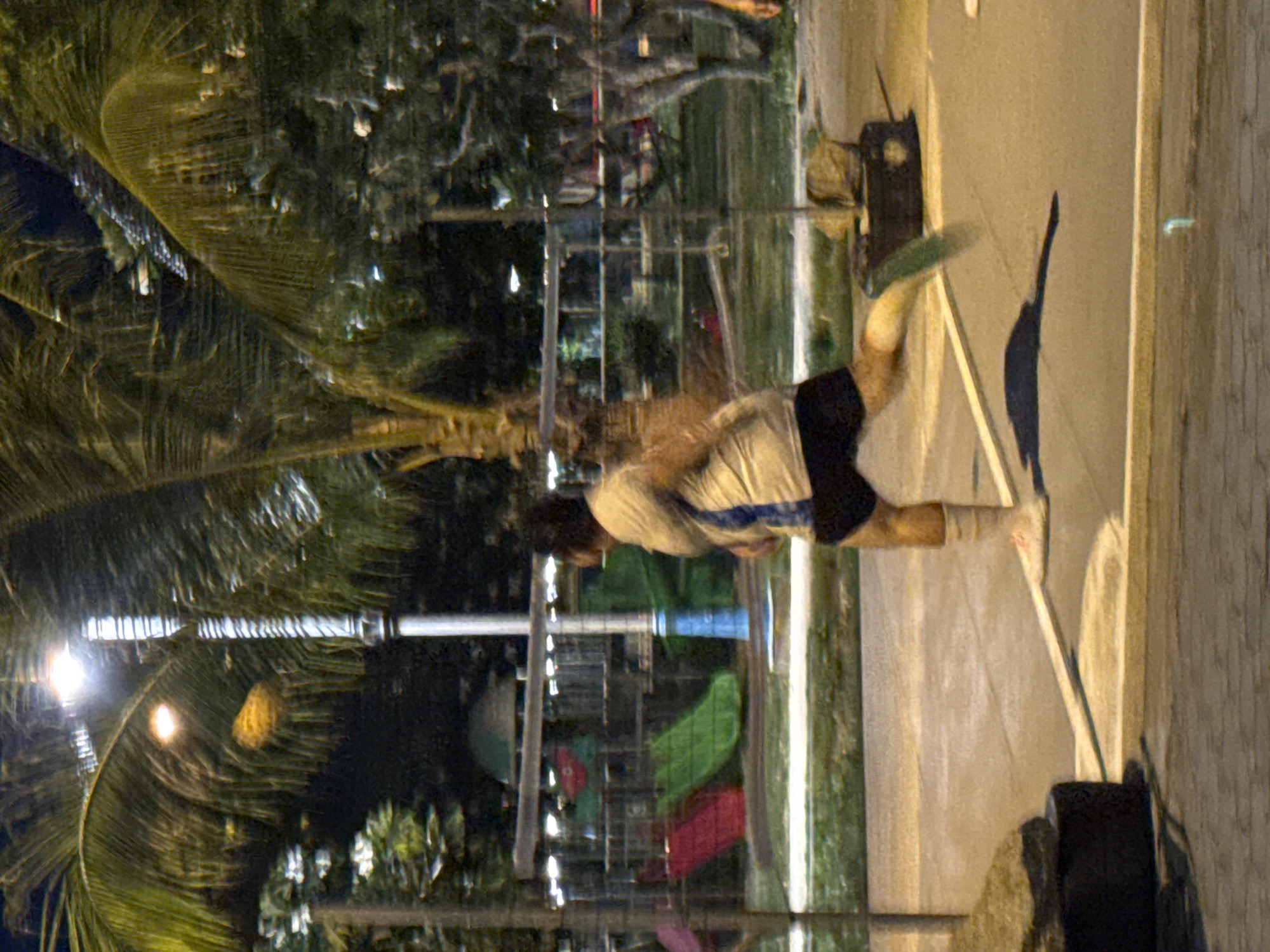 The image depicts a person in motion on a paved surface at night, possibly near a park or recreational area in Koh Phangan, Thailand. A streetlamp with a bright light illuminates the scene, casting shadows. The person, wearing a light-colored shirt with blue accents and dark shorts, is blurred due to movement, suggesting they are running or jumping. Their legs are extended, and one foot is in the air. In the background, there appears to be playground equipment with red and green slides, and palm trees, consistent with a tropical island setting. The overall lighting suggests it is after sunset.