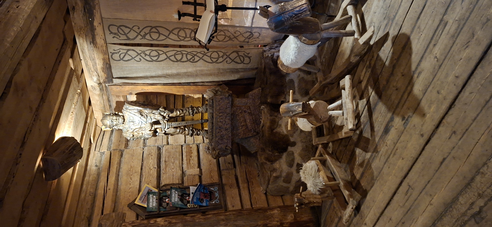 The image depicts an indoor scene within a rustic wooden structure, likely a Viking-themed exhibition or museum, located in Saula, Estonia. Dominating the center is a life-sized, metallic statue of a Viking warrior, complete with helmet, shield, and sword, standing on an intricately carved stone pedestal. The statue is illuminated by a spotlight from above, casting shadows. To the left of the statue, a wooden rack holds several magazines with Viking-themed covers. Behind the statue, a natural stone wall is visible, covered with animal pelts, suggesting a historical or primitive setting. To the right of the statue, a beige fabric tapestry with a dark, intricate knotwork pattern hangs vertically, adding to the Viking aesthetic. In front of the tapestry, a metal stand holds an open book, and a candle in a holder is positioned on the stand. The foreground is occupied by three wooden rocking toys: two stylized horses and a rocking sheep, all appearing handcrafted and rustic. The floor is made of rough-hewn wooden planks. The overall impression is one of a carefully curated historical display, evoking the spirit of the Viking era.