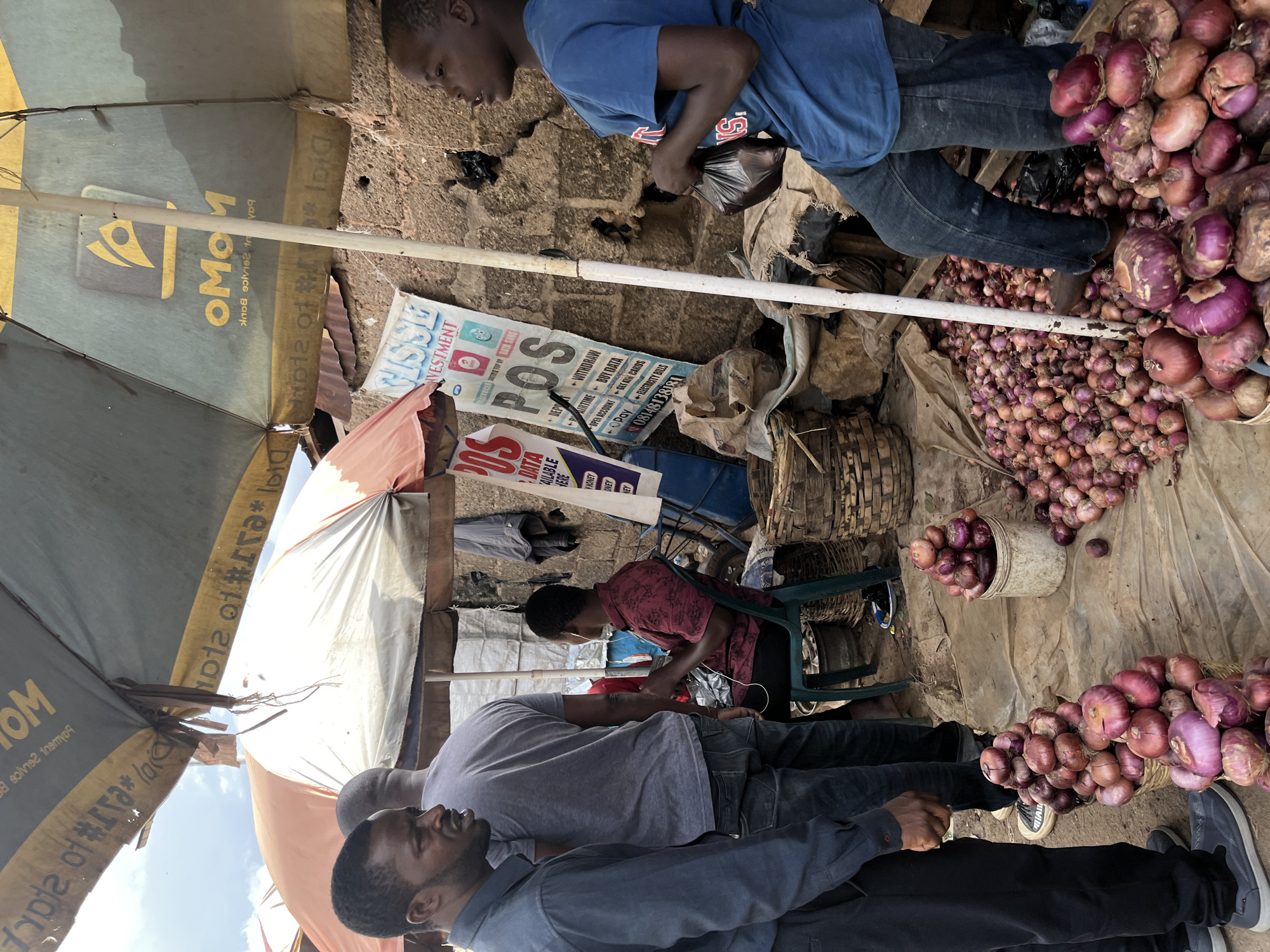 The image depicts a street market scene in Rafin Par, Nigeria. Three men are visible, two standing and one seated. The man on the left, wearing a blue collared shirt and black pants, is facing right, looking at something out of frame. Beside him stands another man in a grey t-shirt and blue jeans, facing away from the camera. To the right, a young boy in a blue t-shirt and jeans is standing, holding a black plastic bag in his right hand and looking to the left. A man in a dark red patterned shirt and black pants sits on a green plastic chair to the left of the boy, wearing white earbuds, and looking to his right.
The market stall is covered by large umbrellas, one dark green with a yellow logo that reads "MoMo" and another orange and white one. Various posters advertising "POS" services are displayed on a rough, textured brick wall. In the foreground, several large piles of red onions are arranged on the ground, some in woven baskets and one in a white bucket. Other baskets and sacks are also visible, along with a black garbage bag. The ground is covered with a textured, tan material. The overall environment suggests a busy, outdoor marketplace.