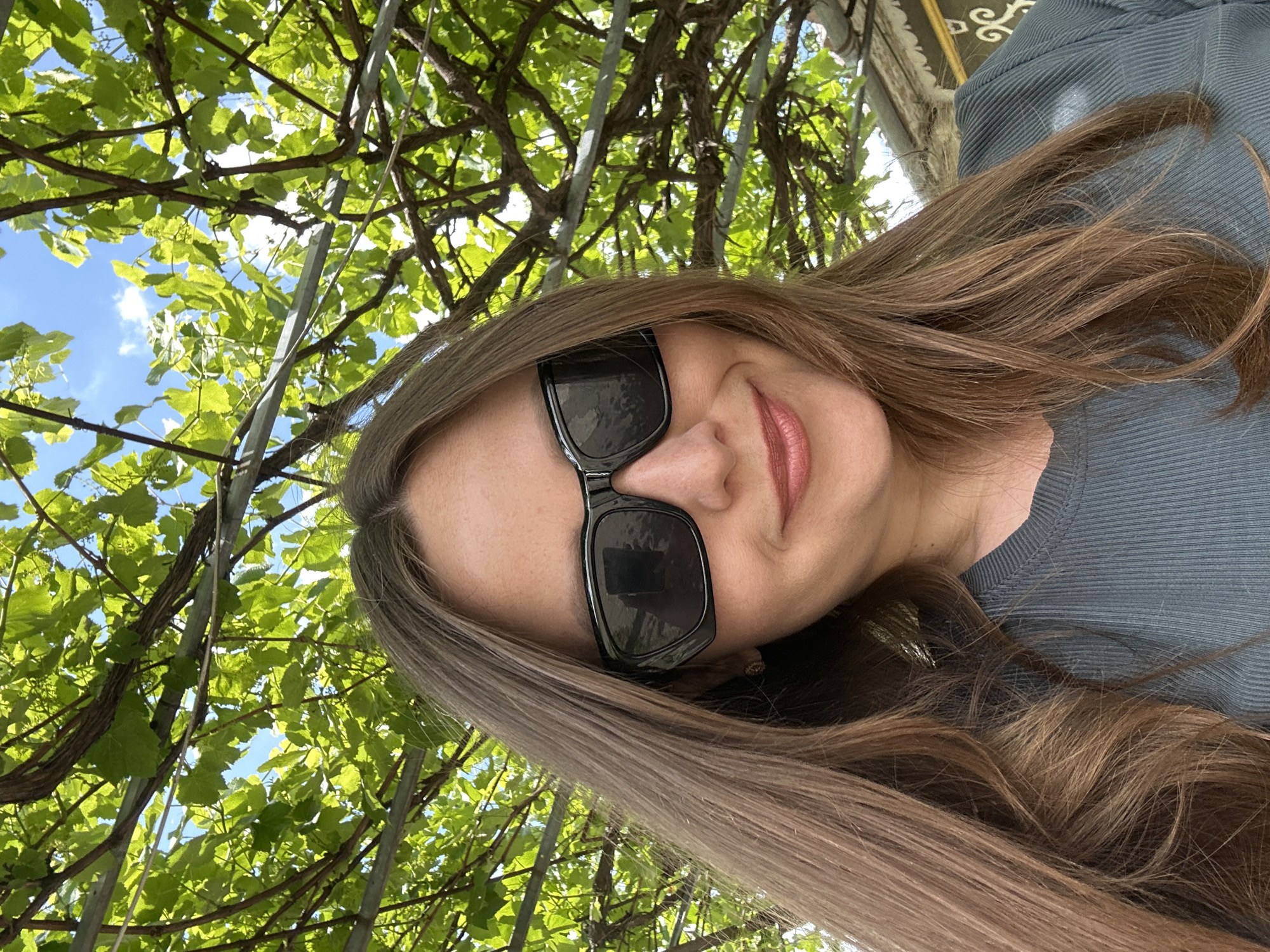 The image is a close-up, slightly low-angle selfie of a woman with long, brown hair and sunglasses. She is smiling and looking at the camera. She is wearing a ribbed, dark grey top. The background is out of focus, but appears to be an outdoor setting with lush green foliage overhead and a clear blue sky visible through the leaves. Metal framing for the foliage is also visible. The overall impression is of a bright, sunny day. There are no specific identifiable landmarks within the image that would suggest the location is in Bila Tserkva, Ukraine.