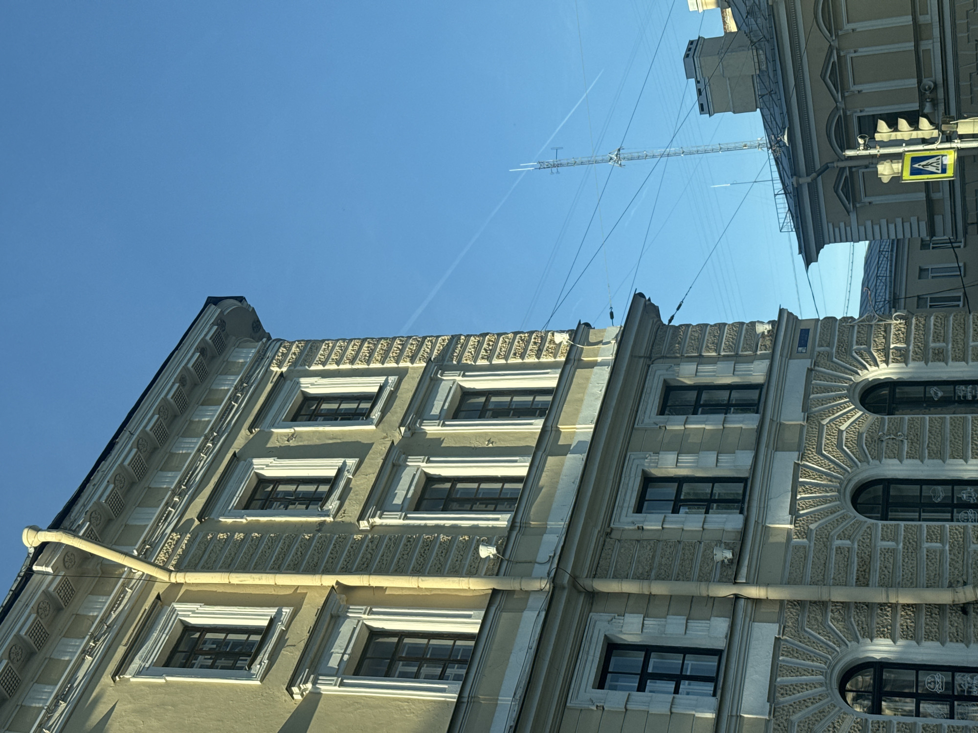 The image shows the side of a yellow building with multiple windows and ornate architectural details, against a clear blue sky. A yellow downspout runs vertically along the side of the building. In the background, a tall radio tower is visible, along with some power lines. To the right of the tower, the roof of another building with a chimney can be seen. A traffic light and a pedestrian crossing sign are also present in the lower right portion of the image. The overall scene depicts an urban environment.