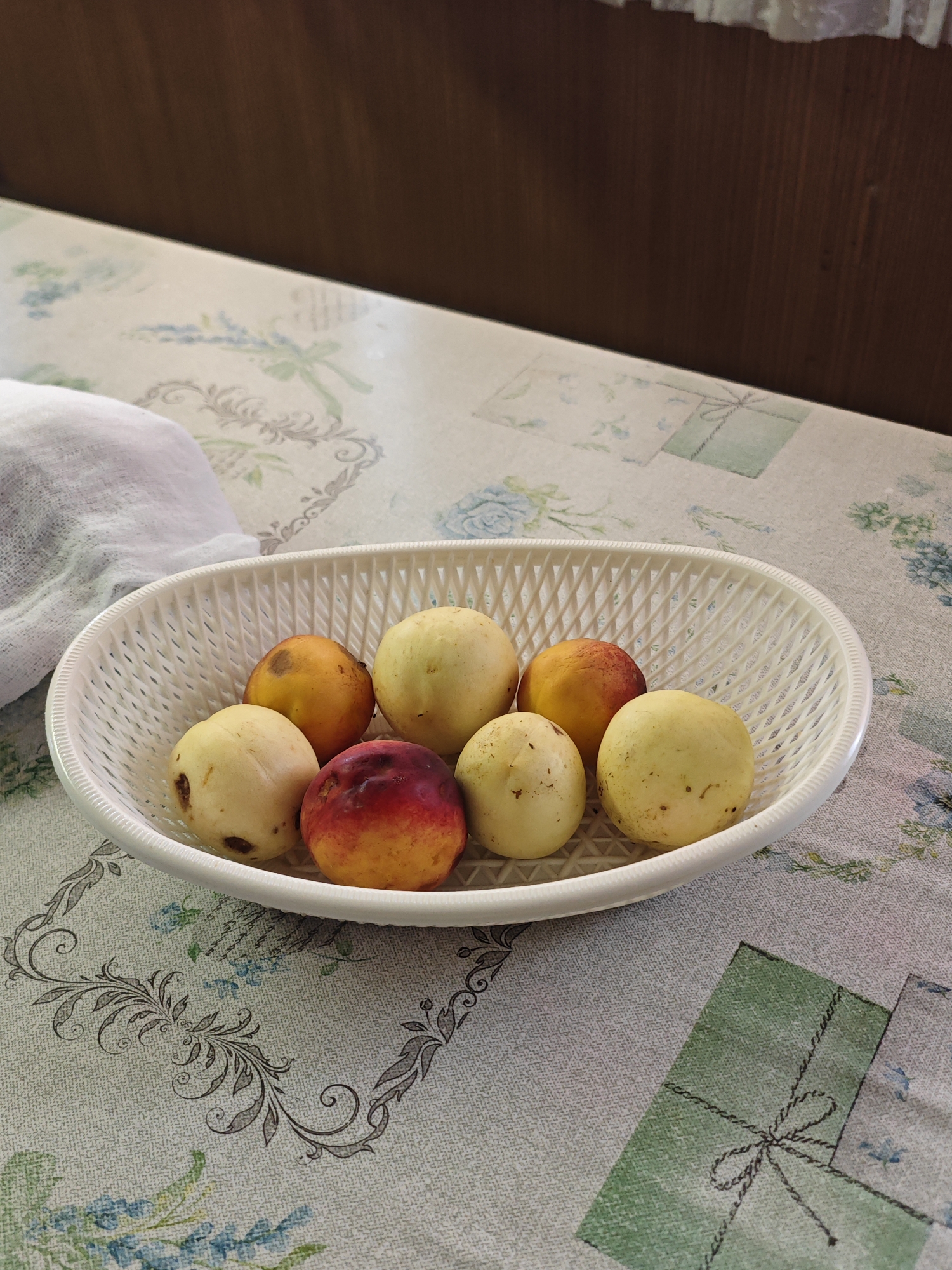 A white plastic basket containing seven assorted fruits sits on a patterned tablecloth. The fruits appear to be peaches or nectarines, with varying shades of yellow, orange, and red, and some showing browning. To the left of the basket, a crumpled white cloth is visible. The tablecloth has a delicate floral pattern with some square elements that resemble wrapped gifts. In the background, a dark wooden surface is present, and a white lace trim is visible at the top edge. The overall setting suggests a domestic indoor environment. The image does not contain any information that would allow for a specific location within the city of Yartsevo, Russia.