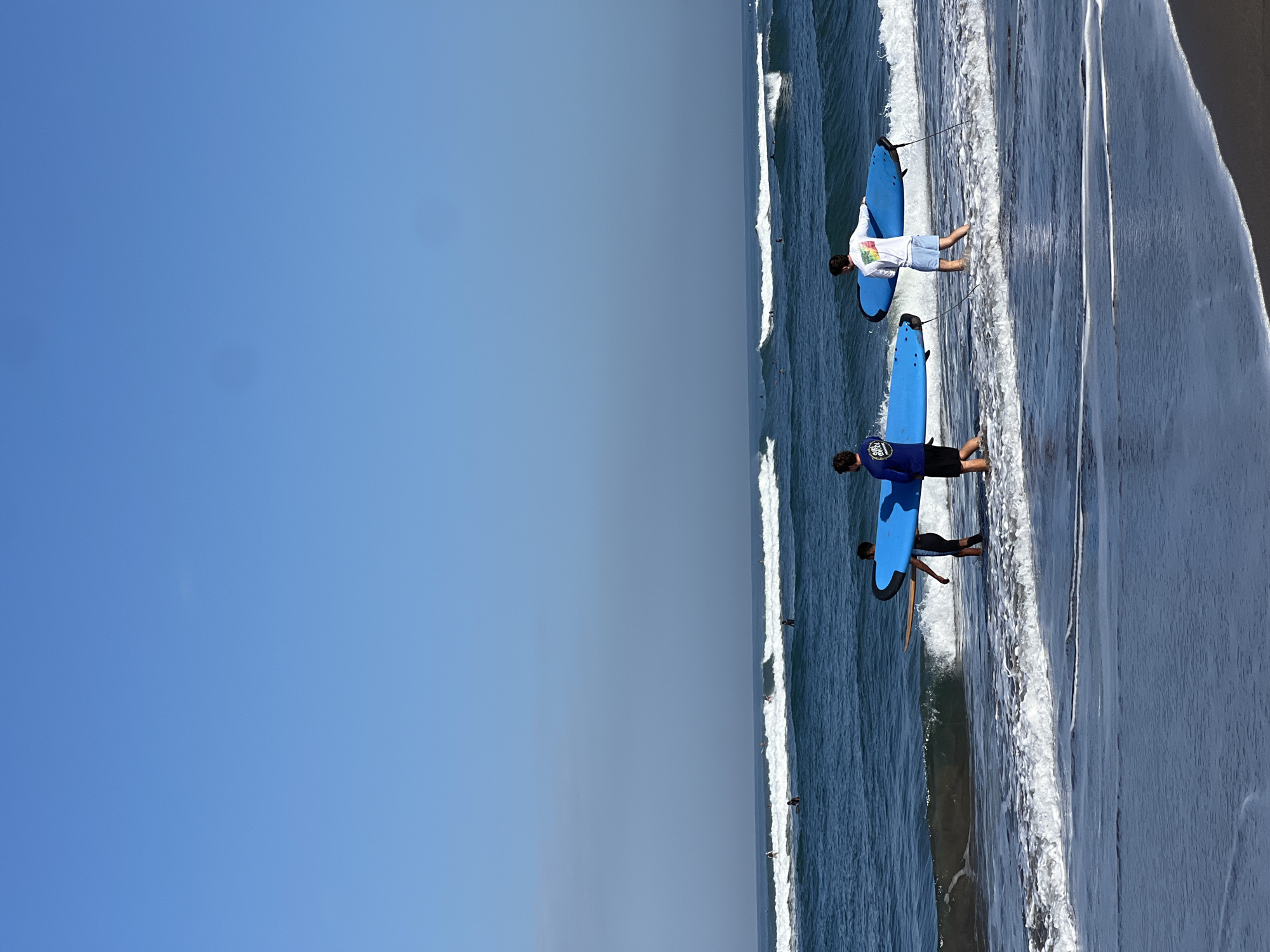 In North Kuta, Indonesia, a clear blue sky stretches over a tranquil ocean where gentle waves roll onto a sandy beach. Three individuals, two young males and a third male, are walking into the shallow water, each carrying a bright blue surfboard. The two individuals in the foreground, closer to the shore, are facing away from the camera, while the third individual, slightly further out, is also facing away. Several other surfers are visible in the distance, scattered across the water, enjoying the waves. The scene captures a peaceful moment of recreational activity on a sunny day.