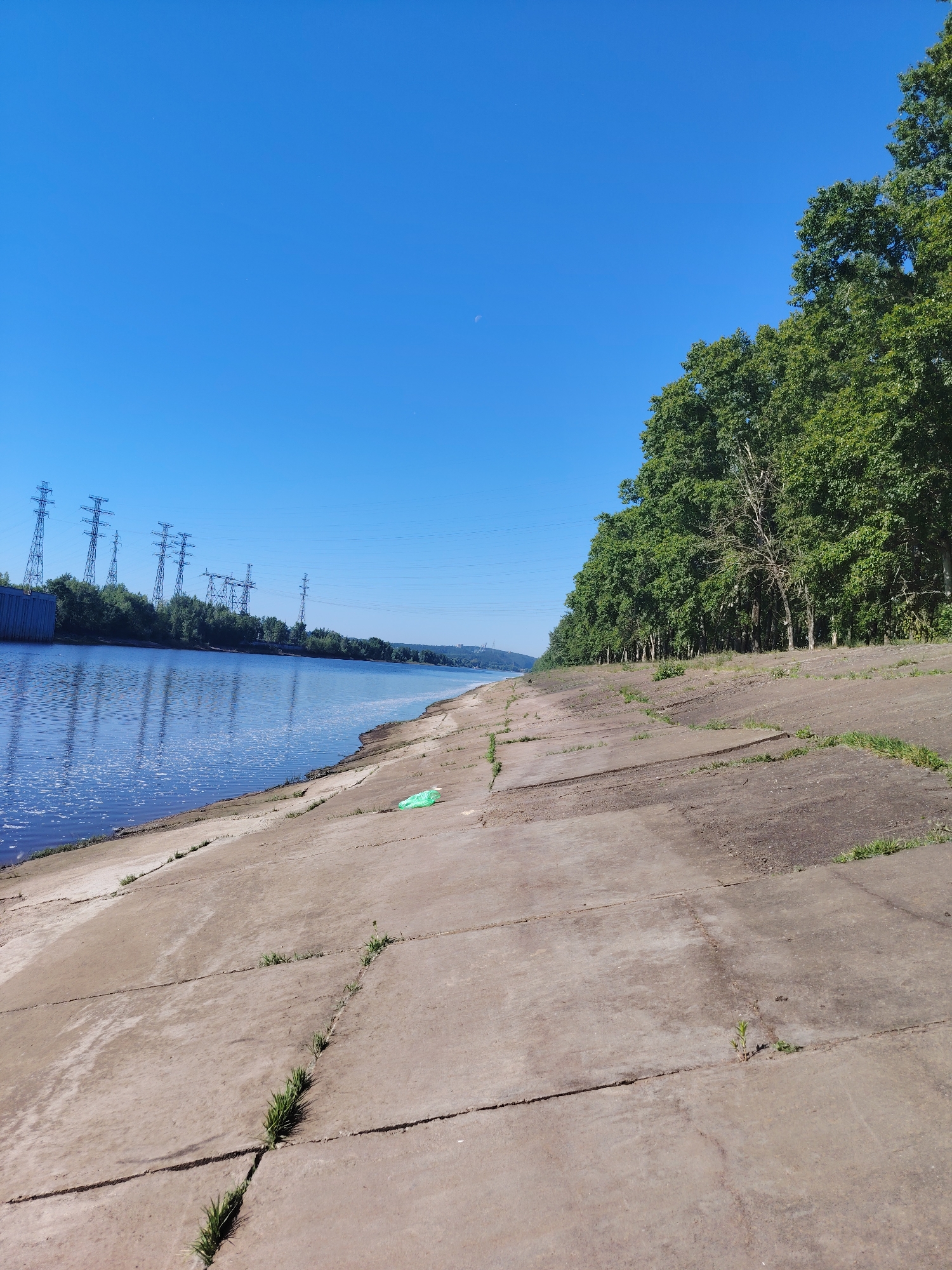 This is an eye-level, wide-angle, outdoor, daytime, horizontal shot of a concrete embankment along a wide river. The sky is a clear, bright blue with a barely visible crescent moon. The river is on the left, reflecting the blue sky and a line of green trees on the opposite bank. Several tall power line towers are visible in the distance on the left side of the river. The concrete embankment in the foreground is made of large, square slabs and is partially covered with dry earth and sparse green grass growing in the cracks. A single, bright green plastic bag lies on the embankment in the middle ground. The embankment slopes upwards from the river to a dense line of green trees. The scene appears to be in the city of Chaikovsky, Russia, likely along the Kama River. There are no people or animals visible in the image.