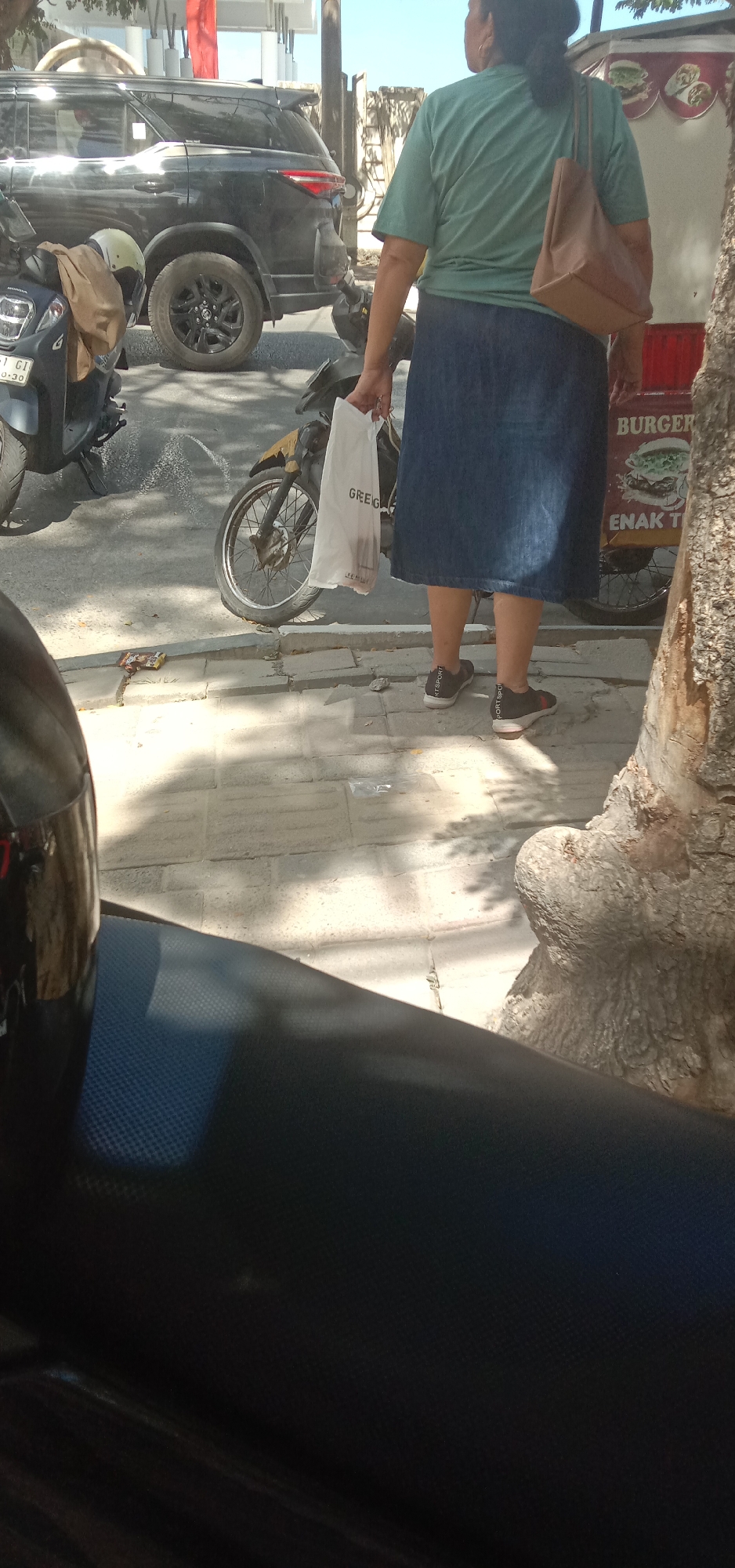 In Kendari, Indonesia, a woman wearing a green t-shirt and a dark denim skirt stands on a sidewalk next to a street. She is holding a white paper bag with "GREG" printed on it and has a brown tote bag slung over her shoulder. Behind her, a black SUV is parked, and two motorcycles are visible. To the right of the woman is a food stall with a sign that reads "BURGER" and "ENAK T." The sidewalk is paved with gray tiles, and shadows from trees fall across it. The scene appears to be a bustling street in the city.