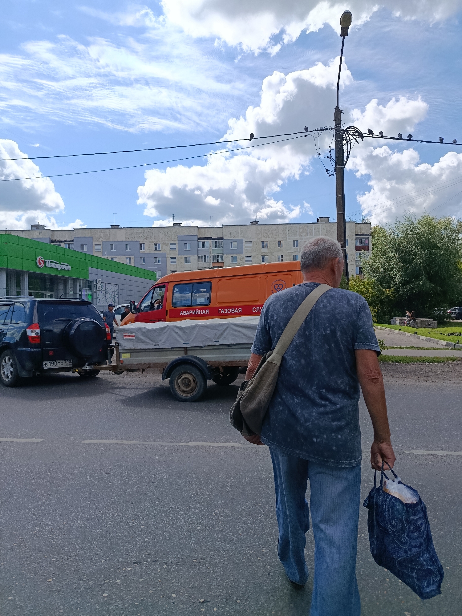 An elderly man walks away from the camera on a paved road in Orekhovo-Zuyevo, Russia. He is wearing a blue, tie-dye t-shirt, blue jeans, and carrying a dark blue patterned bag in his right hand and a grey satchel over his left shoulder. In the background, there is an orange utility van with "АВАРИЙНАЯ ГАЗОВАЯ СЛУЖБА" (Emergency Gas Service) written on its side, hitched to a trailer. A dark colored SUV is parked in front of the van. To the left of the van is a green building with a red "Пятёрочка" logo. The scene takes place on a sunny day with a blue sky and scattered clouds. Birds are perched on power lines in the background.