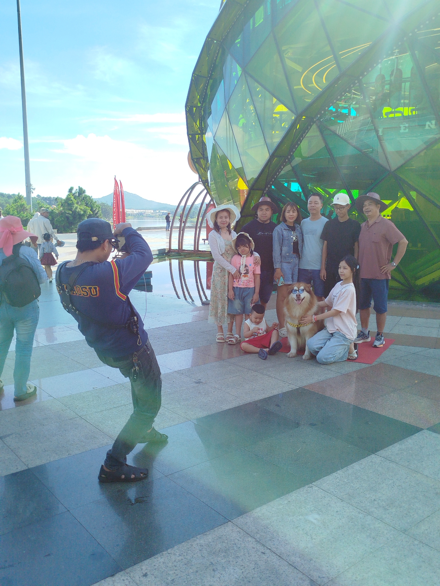 A group of people and a dog are posing for a photo in front of a large, green, geometric building. A man in a blue shirt and a dark cap is taking the photo, bending down to get a good angle. The group includes several adults and two children. One child is sitting on the ground, and another is standing next to the dog. The dog, a large breed with brown and white fur, is wearing sunglasses. The background shows a body of water with a distant mountain and trees. The scene appears to be a public outdoor area, possibly a park or promenade. The architectural style of the building and the general atmosphere suggest a tourist destination in Da Lat, Vietnam.