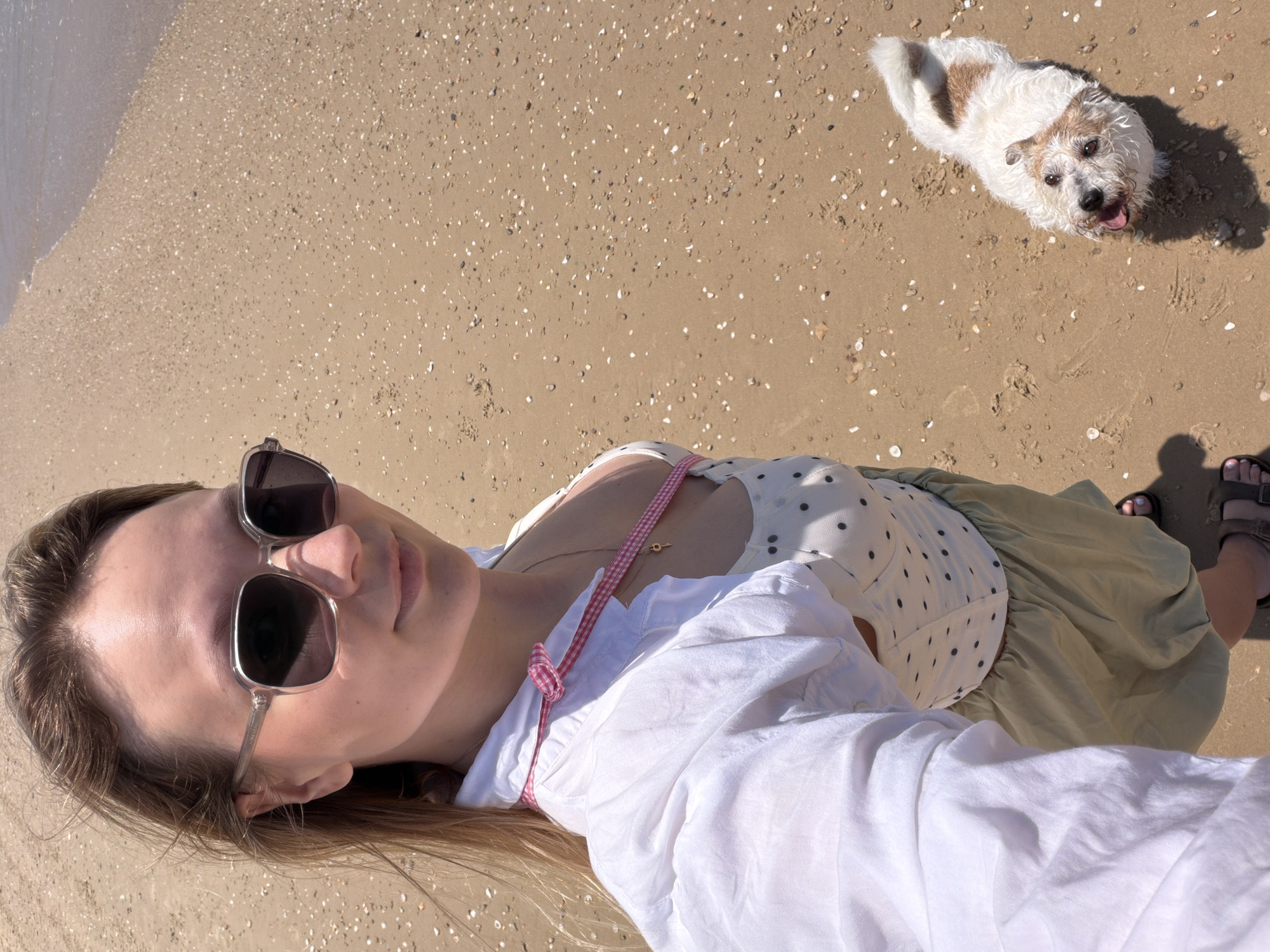 A woman and a dog are on a beach. The woman is lying down, wearing sunglasses, a white top over a polka dot dress, and sandals. She is lying on a sandy beach. A dog is standing near her head. The beach is sandy and appears to be on a sunny day. The scene appears to be taken from the woman’s point of view. Based on the environment of a sandy beach with an ocean visible, the location is possibly on a beach in Tel Aviv, Israel.