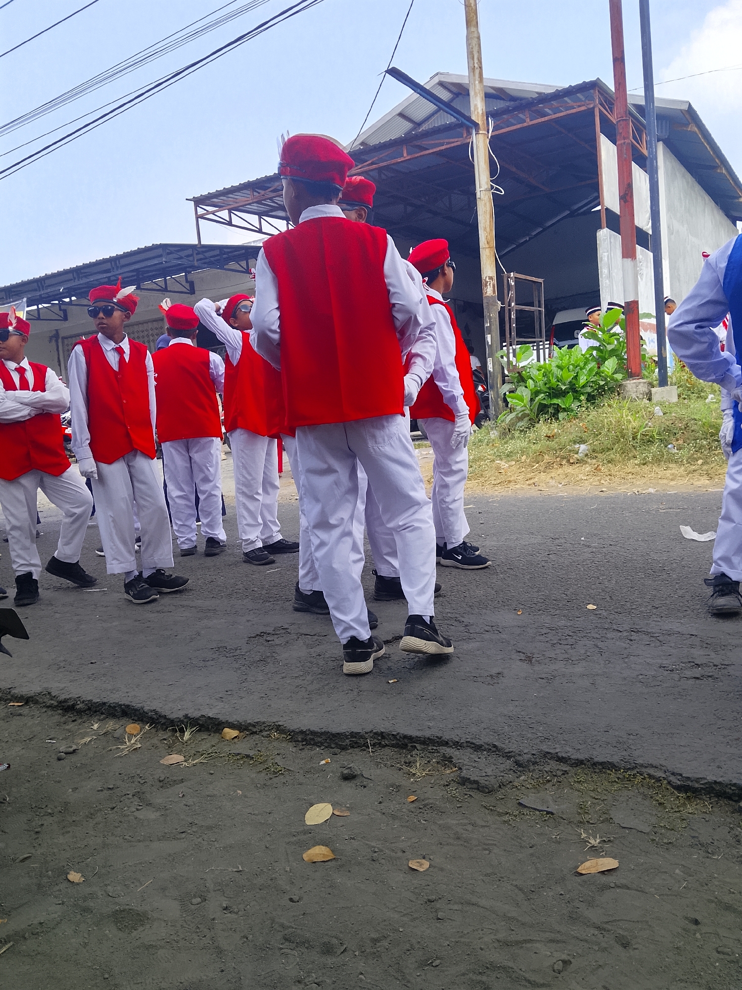 A group of boys in matching red vests, white shirts, and white pants stand in formation on a paved street. Most wear red berets, some adorned with white feathers. The boys appear to be part of a marching band or a similar organized group. In the background, a low-rise building with a corrugated metal roof is visible, alongside a large green bush. The sky is overcast. The scene is likely taking place in Bangsri, Indonesia, as indicated by the reference. The ground is a mix of asphalt and dirt, with some scattered leaves. The overall atmosphere suggests an outdoor event or parade.
