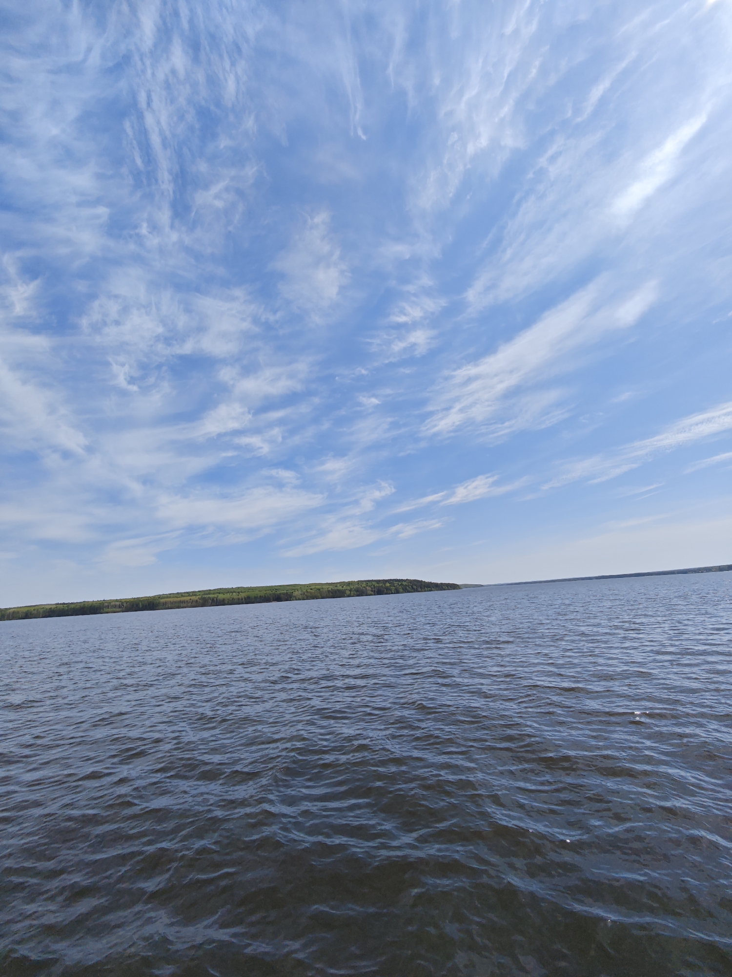 The image depicts a body of water, likely the Votkinsk Reservoir, near Chaikovsky, Russia. The water is dark with gentle waves. A thin strip of land with green trees is visible in the mid-ground, on the far side of the reservoir. The sky is blue with wispy, cirrus clouds streaking across the frame. No people are visible.