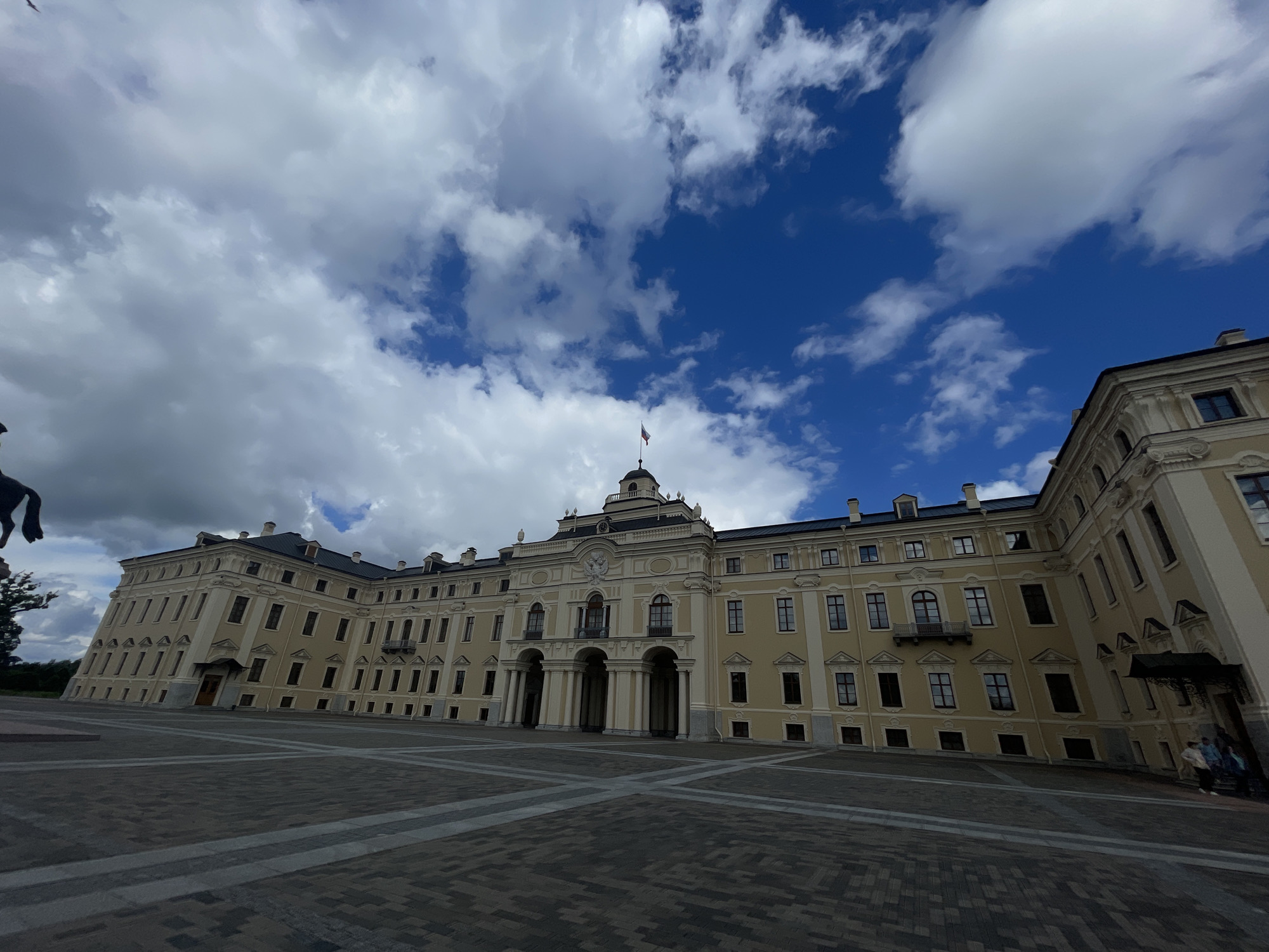 The image captures the Konstantinovsky Palace, located in Strelna, Russia. The grand, pale yellow building features classical architecture with numerous windows, ornate moldings, and a prominent central entrance with arched columns. A flag flies from a dome atop the central section of the palace. In front of the palace is a large, paved courtyard with a geometric pattern. The sky above is a vibrant blue, dotted with dramatic white and gray clouds. The perspective of the photograph is wide-angle, giving a slightly distorted, expansive view of the scene. A few people are visible near the entrance on the right side of the building, and a silhouette of a horse statue is partially visible on the left.