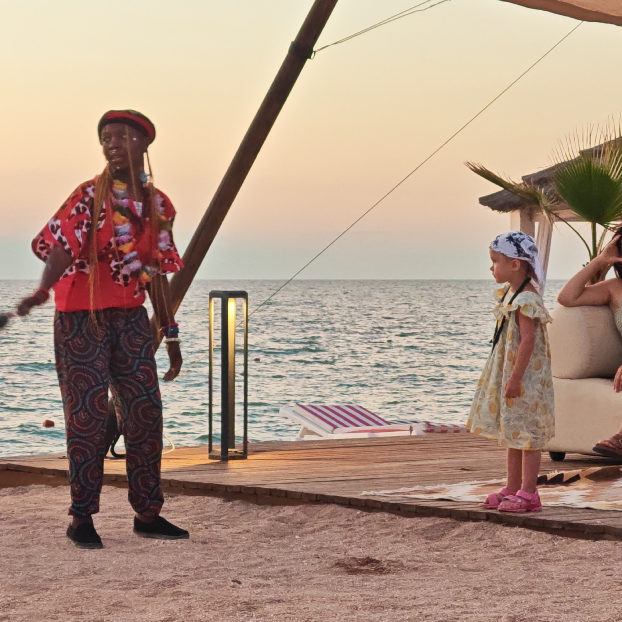 The image depicts an outdoor scene at sunset, likely in Мысовское сельское поселение, Ukraine. In the foreground, a person with dark skin, wearing a red patterned top and patterned pants, stands on a wooden pier. They have beads around their neck and wrists and a red head covering. To their right, a young child wearing a white bandana with palm trees and a light yellow patterned dress stands facing away from the camera. Behind the child, a woman is partially visible, seated on a white couch. The wooden pier extends towards the sea, which is calm and reflects the warm hues of the sunset. A lounge chair with red and white stripes is visible on the pier. A modern lamp post with a warm light is positioned on the left side of the pier. The overall atmosphere is serene and indicative of a beachside gathering or performance.