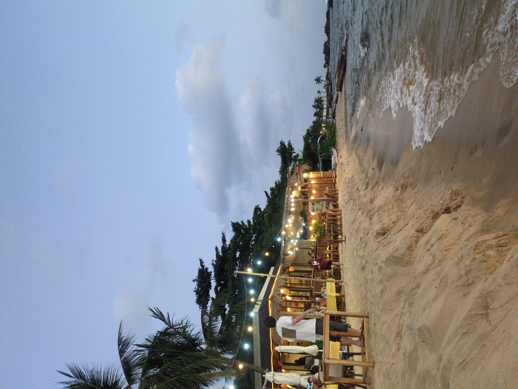 This outdoor scene shows a beachfront establishment in Phú Quốc, Vietnam. The restaurant, adorned with string lights, has wooden tables and chairs arranged on the sand. Several individuals are present, some seated and appearing to be dining, others standing and conversing. The sandy beach extends towards the water, where waves are visible near the shoreline. A line of trees stands between the beach and the restaurant. A palm tree is on the left of the restaurant. The sky is overcast.