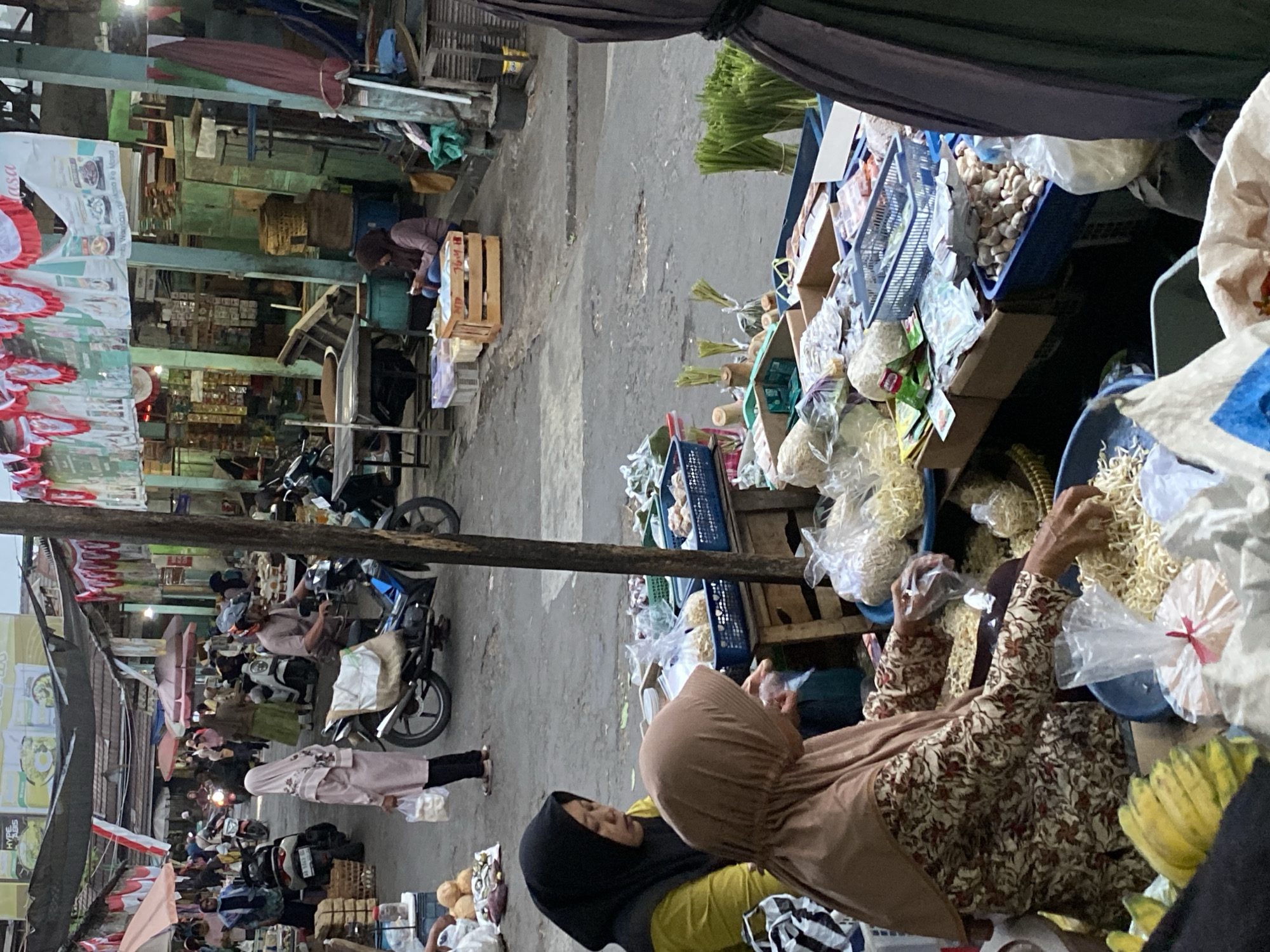 This image depicts a bustling outdoor market scene in Ambarketawang, Indonesia, likely during daytime given the ambient lighting. The foreground focuses on a vendor wearing a patterned brown and beige long-sleeved tunic and a light brown hijab, sorting through a pile of dried noodles in a blue basin. To her left, another vendor, wearing a black hijab and a yellow shirt, observes the noodles. Various packaged goods, baskets of produce like green onions and what appears to be ginger or garlic, and other food items are displayed on wooden stalls.
In the midground, the market extends into a pathway lined with stalls and canopies. Several people are seen browsing or moving through the market. A man wearing a helmet rides a motorcycle with a white sack strapped to the back. A woman in a pink hijab walks with a white plastic bag. Various motorcycles and a scooter are parked in the background. Red and white banners are strung overhead, possibly for a local celebration or event.
The environment is a typical open-air market with a paved ground surface. The architecture visible suggests a more traditional or older part of the city, with simple stall structures and wooden elements. The overall impression is one of lively commerce and daily life in a local Indonesian community.