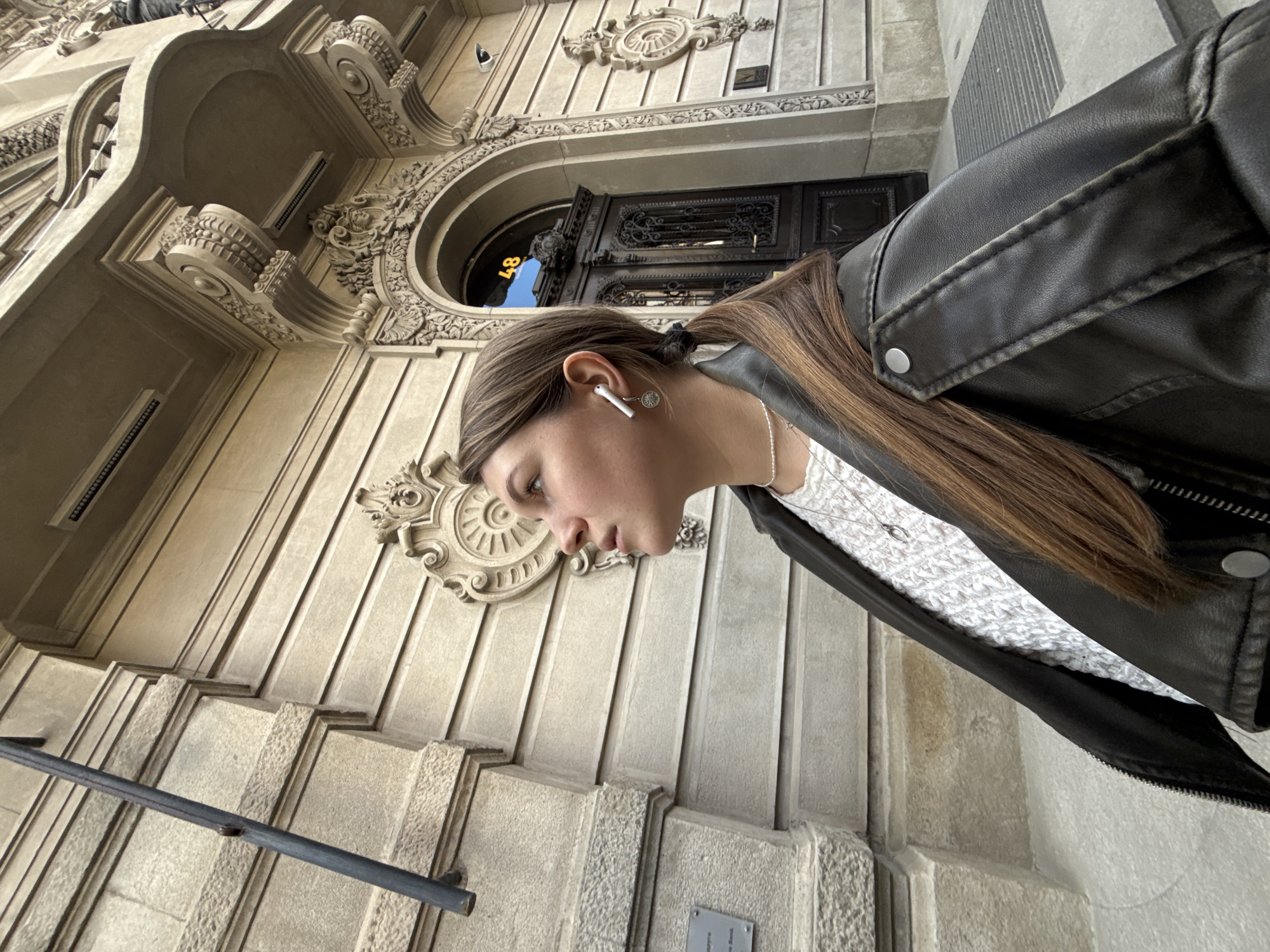 An outdoor, low-angle shot captures a young person partially visible in the foreground against the ornate facade of a building, likely in Belgrade, Serbia. The image is oriented vertically, with the person's head on the left side of the frame, looking towards the top.
The main subject is a young person, likely female, with long, straight brown hair tied back in a ponytail. She wears a black leather jacket over a white, patterned top and a thin silver necklace. A white wireless earbud is visible in her right ear, along with a small, circular silver earring. Her face is seen in profile, with a neutral and somewhat focused expression as she looks forward.
The background features a grand, light-colored stone building with elaborate architectural details. These include highly decorative carvings, moldings, and arched windows. A prominent feature is a large arched entrance with dark, intricately detailed double doors, possibly made of wrought iron or carved wood. The number "48" is clearly reflected in yellow on a surface above the door. Stone steps lead up to the building, flanked by a simple dark metal handrail on the lower left. The facade is adorned with classic architectural elements like rosettes, volutes, and cornices, indicating a building of historical significance. A small, unreadable metallic plaque is affixed to the wall near the bottom right.
The scene takes place during the daytime under clear weather conditions, suggested by the bright, even lighting and lack of shadows. The person appears to be either walking past or standing near the building, possibly listening to audio through her earbud. The overall setting evokes a vibrant European city environment.