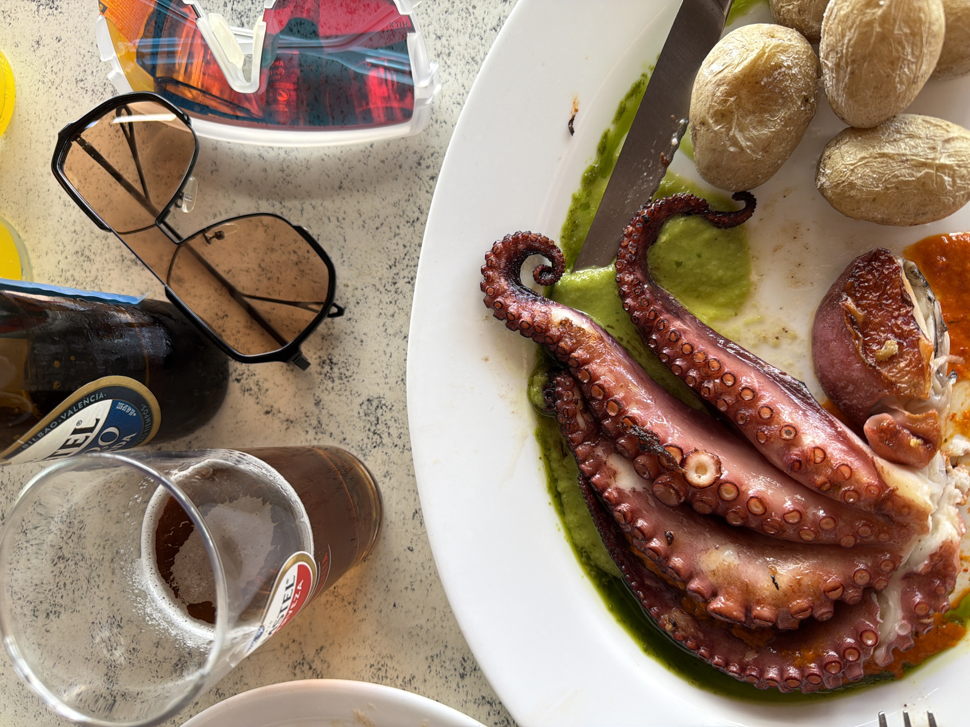 An overhead, close-up shot captures a meal laid out on a light-colored, speckled table surface, likely at a casual outdoor dining setting in Tías, Spain. The central focus is a large serving of grilled octopus tentacles artfully arranged on a white plate. The octopus is accompanied by a vibrant green sauce or puree and a reddish-orange sauce, along with several small, unpeeled, whole potatoes, characteristic of "papas arrugadas" from the Canary Islands, some showing visible salt crystals. A silver knife rests on the plate next to the food.
To the left of the plate, a pair of black-framed sunglasses with dark, reflective lenses is positioned. Behind the sunglasses, a colorful, abstract object, possibly a piece of artwork or a transparent souvenir, reflects various hues including red and orange. Further down, a brown glass beer bottle is visible, with part of its label showing "VALENCIA." Next to the bottle, a glass of beer, partly consumed with foam at the top, is present. The brand name "AMSTEL" is clearly visible in red lettering on the side of the beer glass.
The bright lighting suggests it is daytime. The arrangement of food and drinks implies a relaxed dining experience. No people are visible, and no specific emotions are conveyed directly, but the scene evokes a sense of enjoyment and leisure.