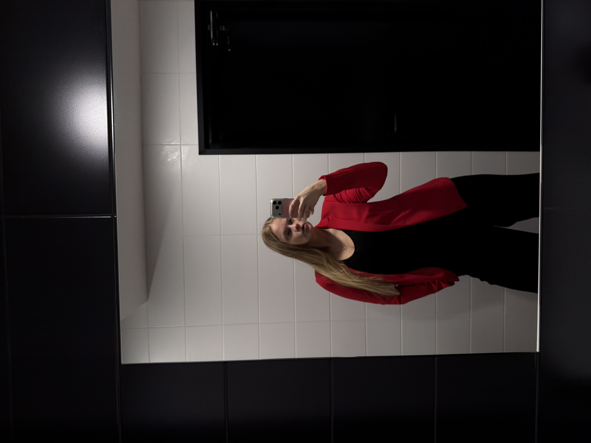 A mirrored image shows a young woman in a black top and red blazer taking a selfie with her smartphone. She is positioned in a bathroom, which has black and white tiled walls and a black cabinet or door frame. The reflection of a rectangular object with a handle can be seen above her head, likely part of the bathroom fittings. The environment appears modern and well-lit. There's no evidence of the city Izhevsk, Russia.