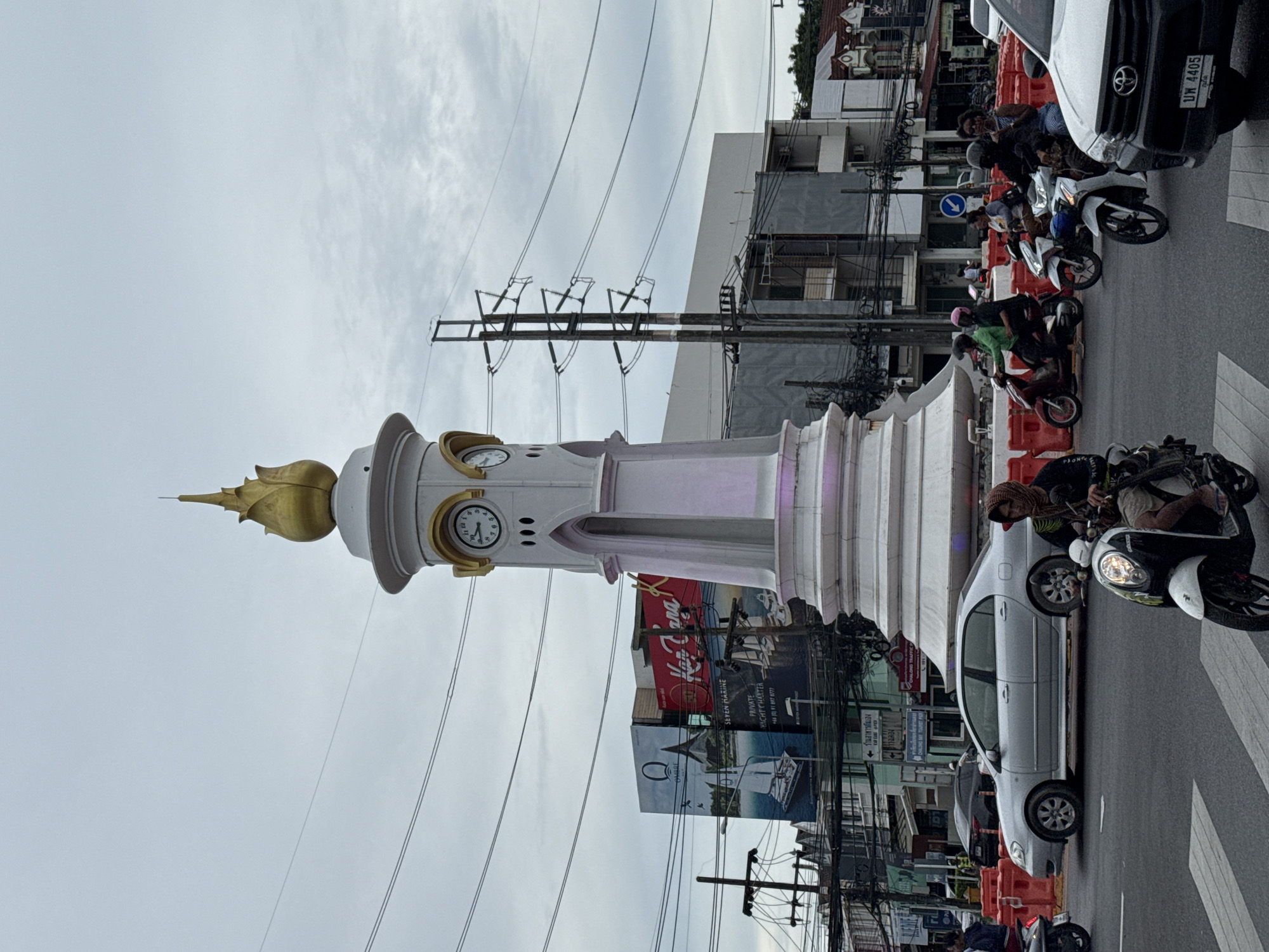 The image depicts a street scene in Chalong, Thailand, dominated by a tall, white clock tower topped with a golden, lotus-like ornament. The tower has clock faces on its upper section. Surrounding the tower are various buildings with billboards, some displaying advertisements for "Ombre" and "Kan Tiara." Electrical wires crisscross the sky above the street.
The foreground and midground are filled with traffic, primarily motorcycles and cars. Several people are visible riding motorcycles. One person on a white scooter in the lower foreground is wearing a headscarf and shorts. Another scooter behind it has two people, one wearing a green jacket and helmet, and the other a pink helmet. Further back, more motorcycles and a silver sedan are on the road. A white SUV is partially visible on the right side of the frame. The road has pedestrian crossing markings. Orange traffic barriers are present along the side of the road. The sky is overcast, indicating a cloudy day. The overall impression is a busy urban intersection in a tropical climate.