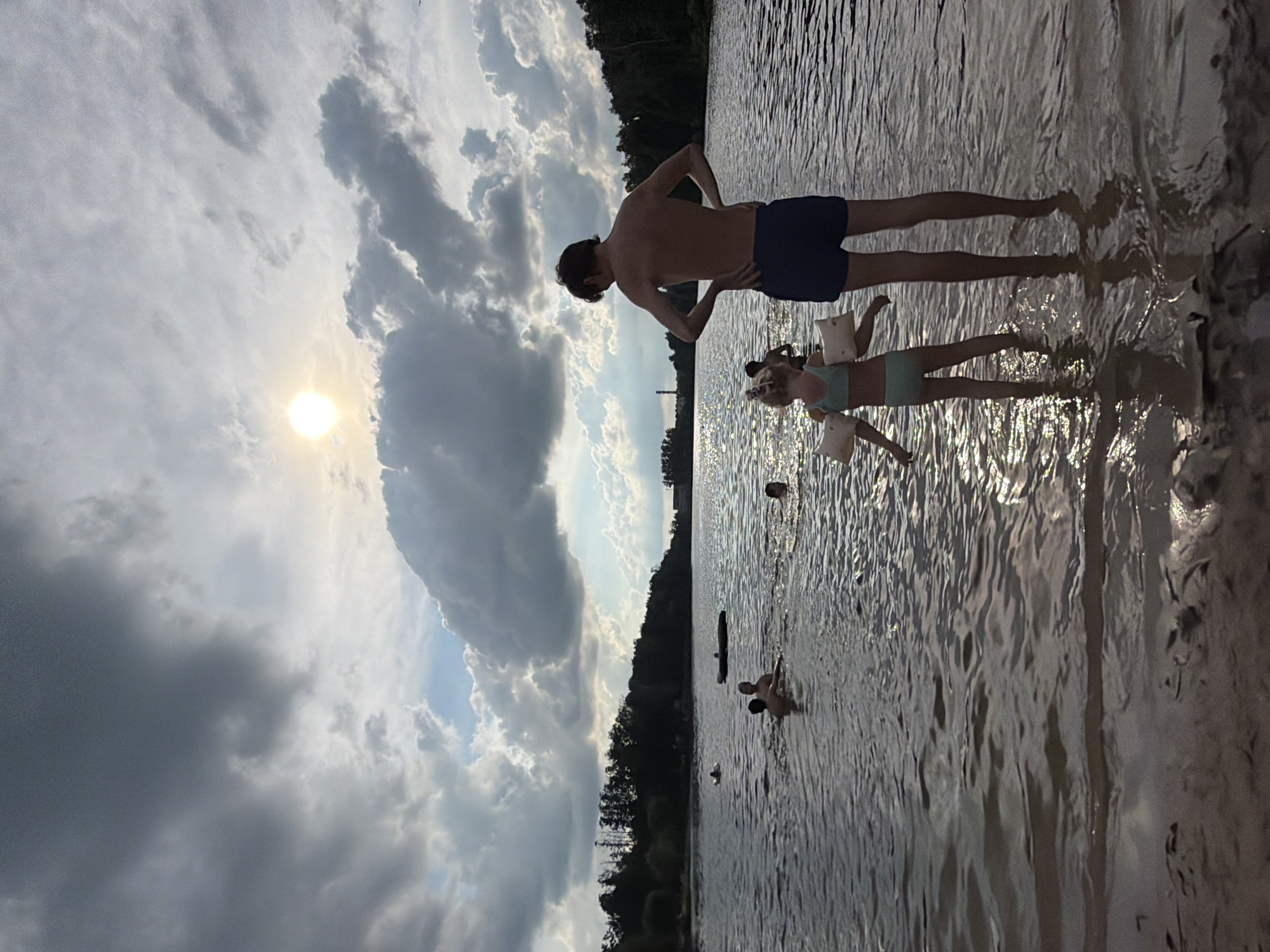 This is a landscape-oriented, low-angle shot of a group of people in a body of water, likely a lake or river, near a wooded shoreline. The sun is setting, casting a warm glow on the water and creating dramatic clouds in the sky.
In the foreground, a man stands with his back to the camera, hands on his hips, wearing dark blue swim trunks. To his right, a young girl in a light blue bikini with inflatable armbands is lying on her back in the water. Further out in the water, two more individuals are partially submerged. The water is calm with ripples reflecting the light.
The background features a dense forest line against a cloudy sky. The sun is low on the horizon, peeking through the clouds, suggesting it is late afternoon or early evening. The overall mood is relaxed and summery. No text is visible in the image.