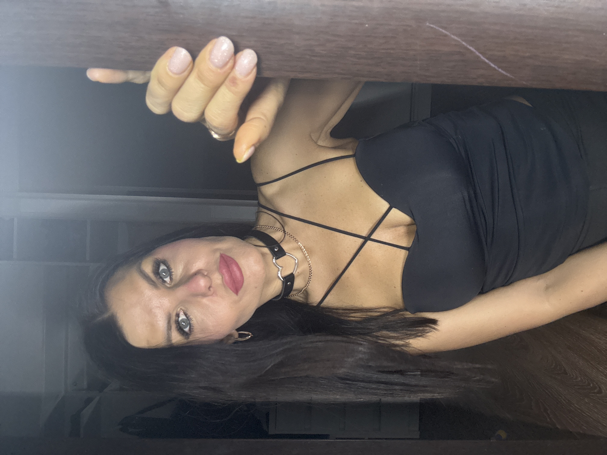 The image shows a woman in a black top, with dark hair, a heart choker necklace, and noticeable eye makeup and lipstick. She is looking at the camera, holding onto a wooden ledge or frame. The environment is dimly lit. There is no information to suggest the location is in Chaikovsky, Russia.