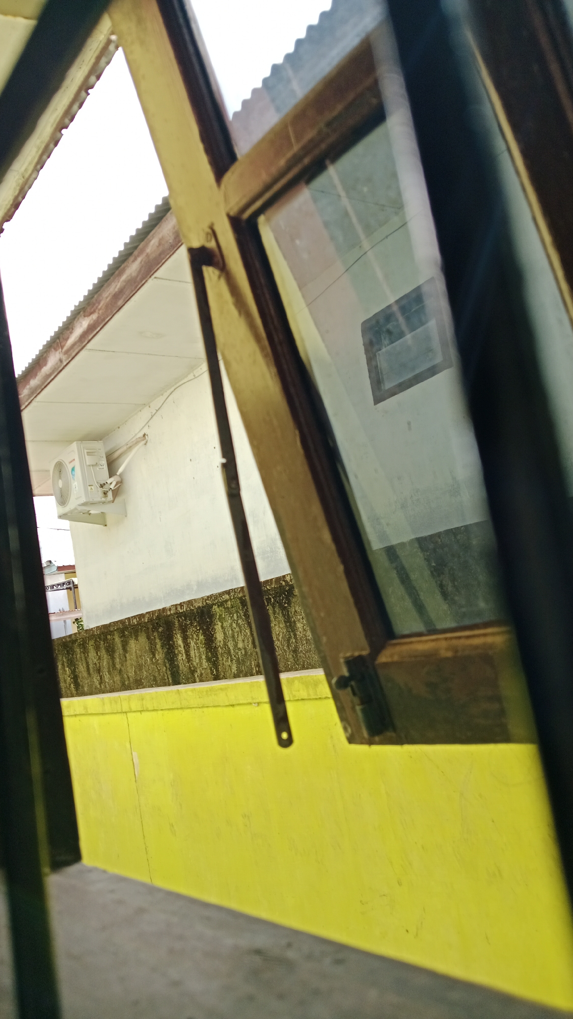 The image is an exterior view from a window in Lhokseumawe, Indonesia. Through the open window, a white air conditioning unit is visible mounted on a light-colored wall. Below the wall, there is a bright yellow painted surface. The foreground shows the wooden frame of the window and a metallic hinge. The sky above is bright, and a corrugated metal roof is visible. Reflections on the window glass obscure some details of the interior.