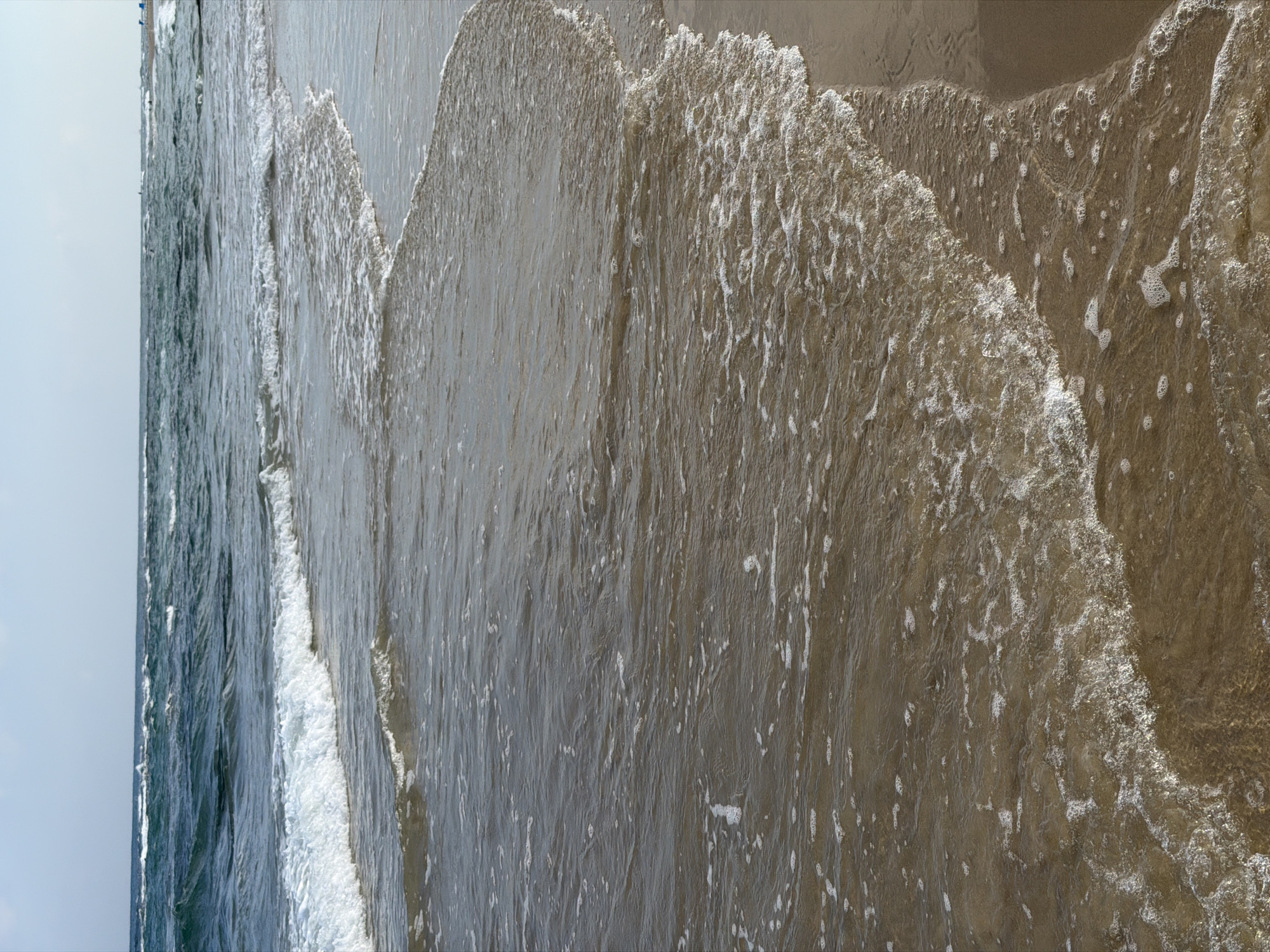 The image captures a view of the Mediterranean Sea from the sandy shore in Bat Yam, Israel. Gentle waves are breaking and receding on the beach, creating foamy white edges and ripples on the shallow water. The water is a deep teal color further out, contrasting with the sandy brown of the wet and dry sand. The sky is a pale blue with some thin clouds. In the distance, faint silhouettes of people can be seen on the beach, some possibly standing in the water. The scene is peaceful and natural, depicting a typical coastal environment.