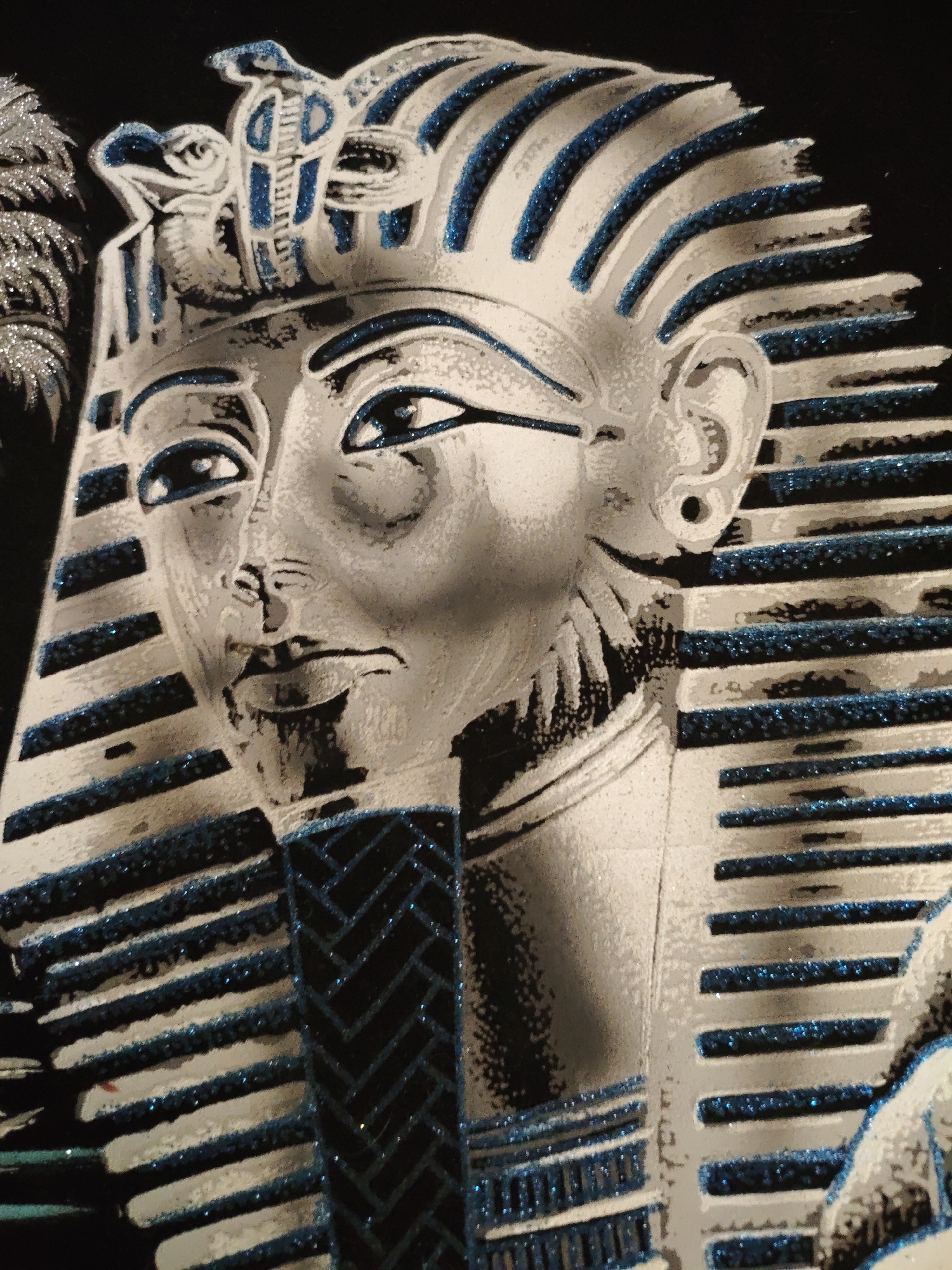 This image depicts a stylized, possibly artificial, representation of an Egyptian pharaoh, likely Tutankhamun, as depicted in his burial mask. The mask is silver or grey with blue accents, possibly glitter, highlighting the contours. The headdress and beard are striped with horizontal blue bands. The eyes are lined with dark blue. The representation is set against a dark background. There is no information indicating the location of the image is in Боровково, Russia.