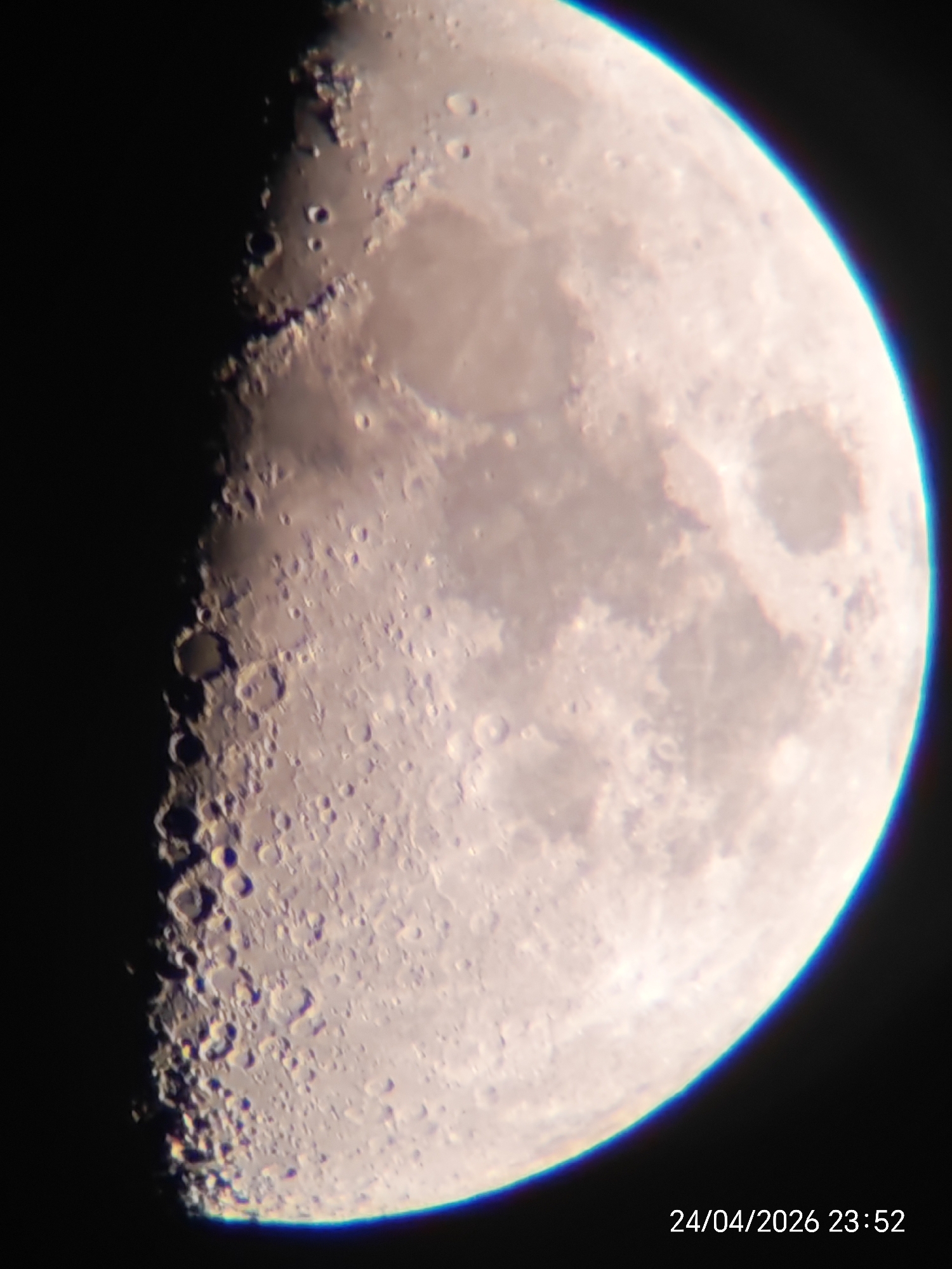 The image presents a detailed close-up of the Moon, illuminated against the dark void of space. Approximately half of the lunar surface is visible, showcasing a prominent terminator running vertically along the left side, separating the sunlit portion from the unlit area.
The illuminated surface reveals a rugged landscape characterized by numerous impact craters of various sizes. Along the terminator, the low angle of sunlight casts long, distinct shadows within these craters, highlighting their depth and sharp relief. The brighter, heavily cratered highlands are clearly discernible across the face of the Moon. Smoother, darker areas, indicative of lunar maria, are also visible, particularly towards the right side of the illuminated disk.
A noticeable optical effect, a faint blue and purple chromatic aberration, is present along the brightly lit limb of the Moon, especially visible on the upper and lower right edges where the lunar disk meets the black sky. This suggests the use of telescopic equipment for capturing the image. The Moon's surface displays natural variations in tones, ranging from light grey to subtle browns.
In the bottom right corner of the image, white text displays the date and time: "24/04/2026 23:52". The photograph captures a moment of astronomical observation, focusing on the intricate details of the Moon's surface during a crescent or gibbous phase.