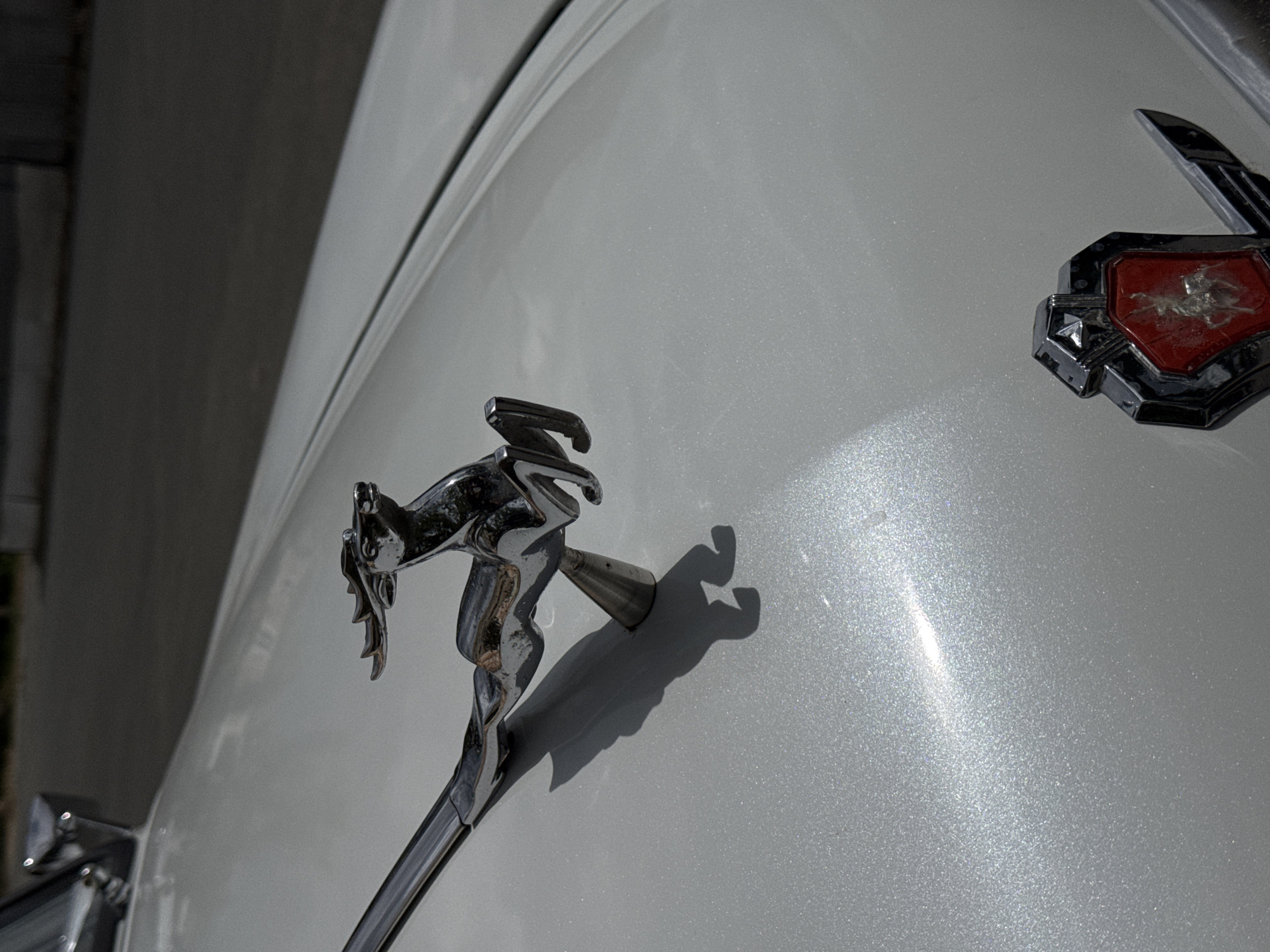 The image presents a detailed, high-angle view of the hood of a car, located in Unknown, Unknown. The car's hood is finished in a metallic light gray or silver paint, which reflects sunlight with numerous small, glittering flakes.
Prominently featured is a chrome-plated hood ornament depicting a stylized, leaping gazelle or antelope-like figure. The ornament is highly reflective, showing distortions of the surroundings, and is mounted on a conical base, which attaches to a polished chrome strip extending towards the front of the vehicle. Strong, direct sunlight illuminates the ornament from the upper left, casting a sharp, dark shadow of the animal onto the car's surface.
In the lower right corner, a dark, angular, shield-shaped emblem is partially visible. It encases a central, red insert that shows significant distress, characterized by multiple scratches and marring, which obscure a faint, lighter-colored figure within. A portion of a dark, stylized wing-like element is visible to the right of the emblem.
The background is uniformly dark and out of focus, suggesting a distant road surface or structure under bright daylight conditions.