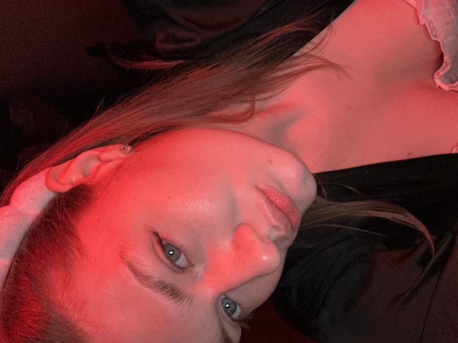A woman with fair skin is angled towards the camera, illuminated by a stark red light. Her blue eyes are visible, with light brown hair falling softly. She wears a delicate earring and what appears to be a sheer, ruffled garment. A dark fabric creates a contrasting background, possibly clothing or a covering. The lighting accentuates facial features, creating a striking, stylized portrait.