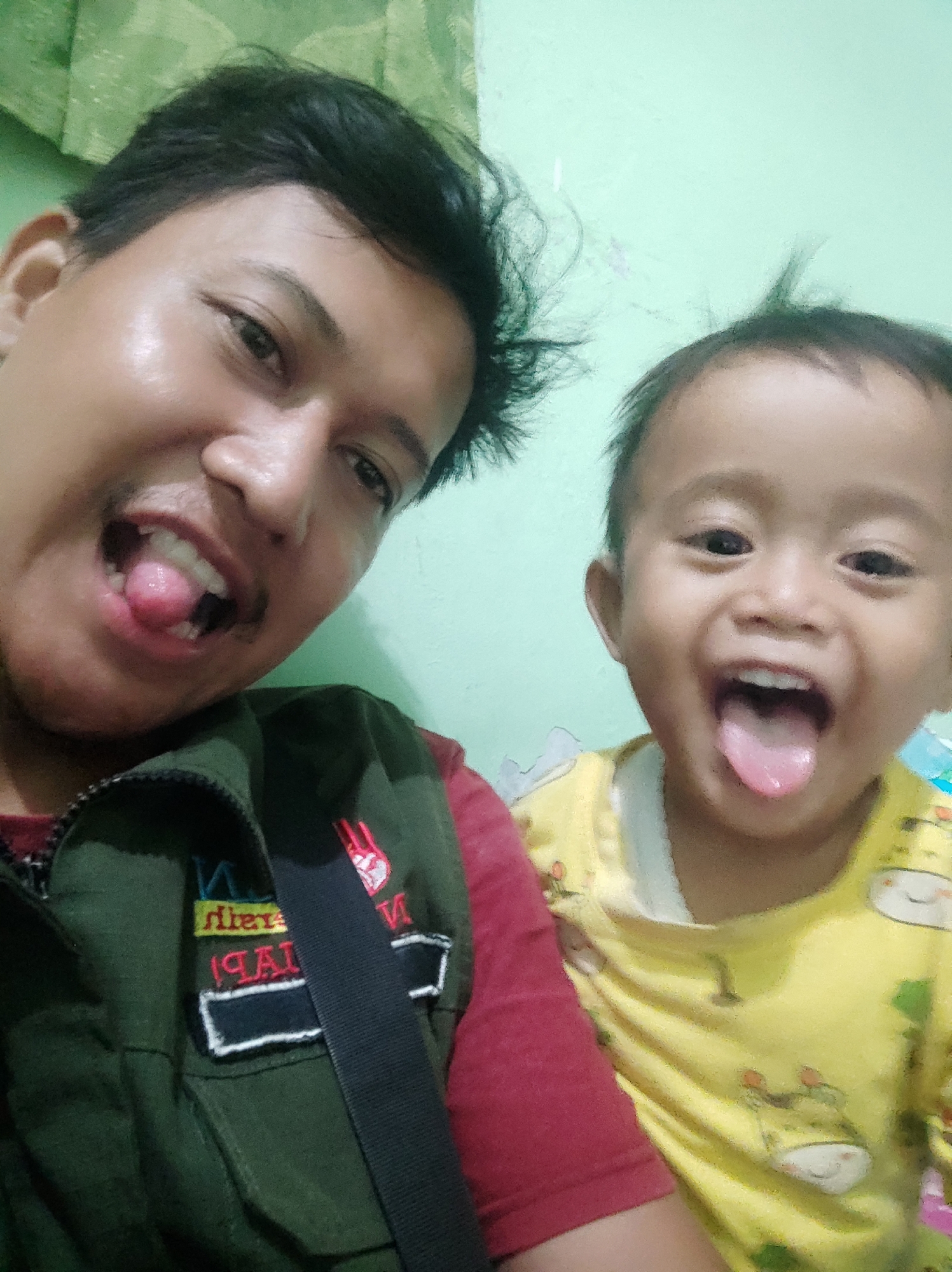 A selfie shows a man and a young child with their tongues sticking out, looking at the camera. The man, on the left, has dark hair and is wearing a dark green vest over a red shirt. The child, on the right, has dark hair and is wearing a yellow shirt with cartoon animal prints. The background is a light green wall. The image appears to have been taken indoors.