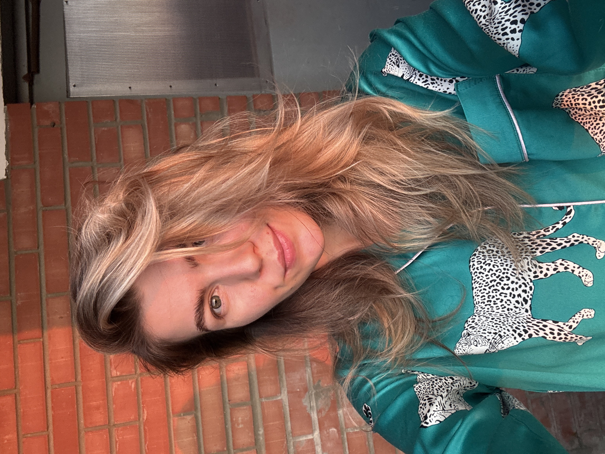 An outdoor, waist-up medium shot shows a woman with blonde hair and fair skin. She is looking directly at the camera with a slight smile. Her hair is long and wavy, with strands falling forward and to the sides of her face. She has on a teal-colored silk pajama top with a leopard print. The background consists of a red brick wall to the left and a gray metal door on the right. The lighting suggests it is late afternoon or early evening, with warm sunlight illuminating the woman and the brick wall.