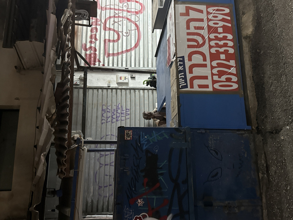 The image depicts an urban scene at night or under low-light conditions, likely an alley or market area in Tel Aviv, Israel, identifiable by Hebrew script and phone number on signage.
The central foreground features a prominent blue-painted counter-like structure, partially covered with cardboard on its top surface. A large white sign with bold red Hebrew text "לְהַשְׂכָּרָה" (For Rent) and the phone number "0522-333-990" is affixed to the front of this blue structure, secured by clear plastic. To the left of this counter is a taller, blue, rectangular utility box or closed stall section, heavily covered in graffiti including a Star of David, abstract shapes, and a stylized face with large eyes and jagged teeth near the bottom.
Behind the blue counter, a single individual, appearing to be an older person with short, light-colored hair, is seated. This individual is seen from the chest up, leaning slightly forward with hands near their face, seemingly in a state of rest or contemplation. The person is wearing a light-colored top.
The background consists of multiple closed metallic roller shutters, indicative of secured shops or market stalls. These shutters are covered in various graffiti, including red script and abstract shapes on the rightmost shutter, and purple/light blue script or shapes on the central shutter. A small white electrical box is mounted on the wall between two shutters on the right. Above the scene, a makeshift canopy composed of several layers of corrugated metal sheets, some appearing damaged or dislodged, provides overhead cover. The ground surface is dark asphalt or concrete, showing minor cracks. Illumination is dim, with artificial light sources casting soft shadows.