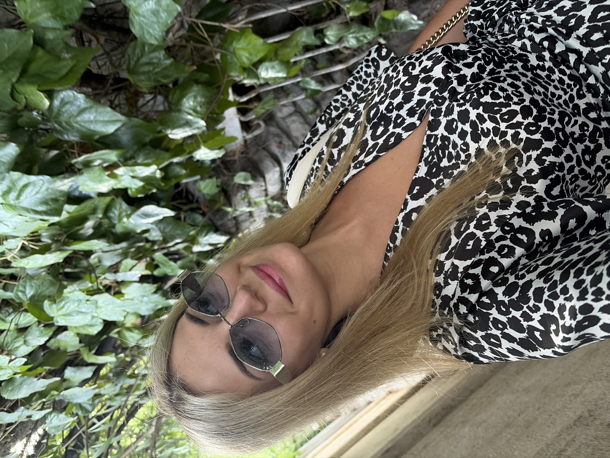In Budva, Montenegro, a blonde woman with blue eyes and sunglasses is seen posing for a selfie in front of a stone wall overgrown with green ivy. She is wearing a black and white leopard-print dress and a gold chain purse is draped over her shoulder. The lighting suggests it is daytime, and the woman is smiling.