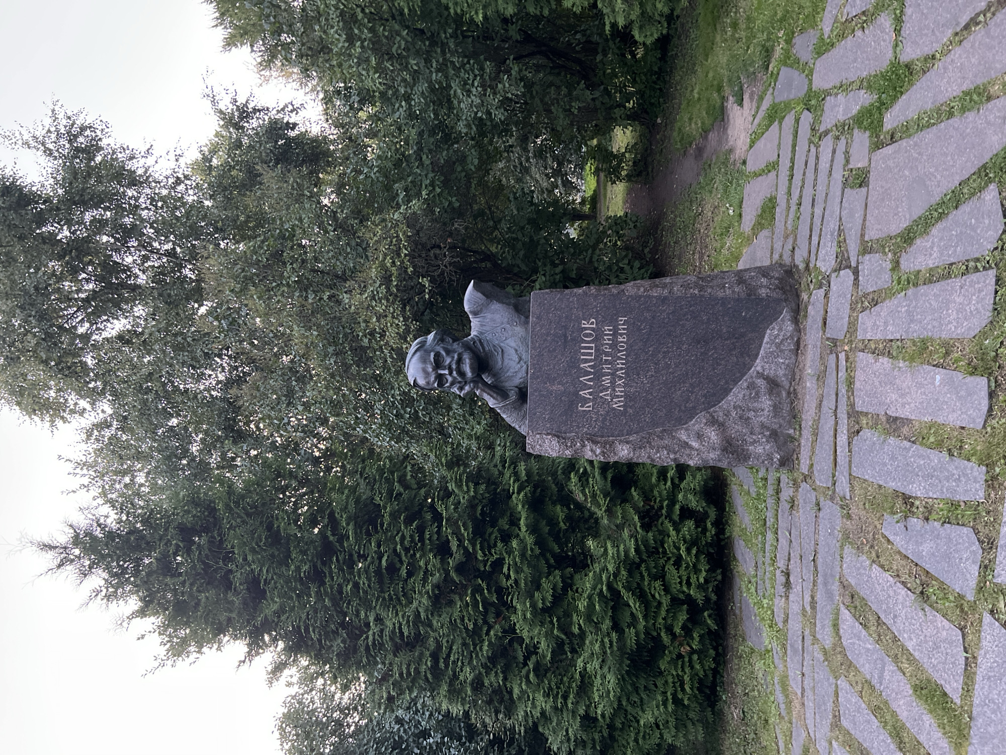 A bronze bust of a man with a beard and mustache rests on a tall, rough-hewn stone pedestal. The bust depicts the man with his chin resting on his left hand, his elbow supported by his right hand. Inscribed on the stone pedestal in Cyrillic script are the words "БАЛАШОВ ДМИТРИЙ МИХАЙЛОВИЧ." The sculpture is situated outdoors, on a pathway made of large, irregular grey stone slabs interspersed with grass. Behind the sculpture, a path winds through a dense green forest with tall trees and evergreens. The lighting suggests it is either early morning or late afternoon. This memorial is located in the city of Veliky Novgorod, Russia, specifically in the Demyansky District on the territory of the memorial complex "Demyansky Pocket."