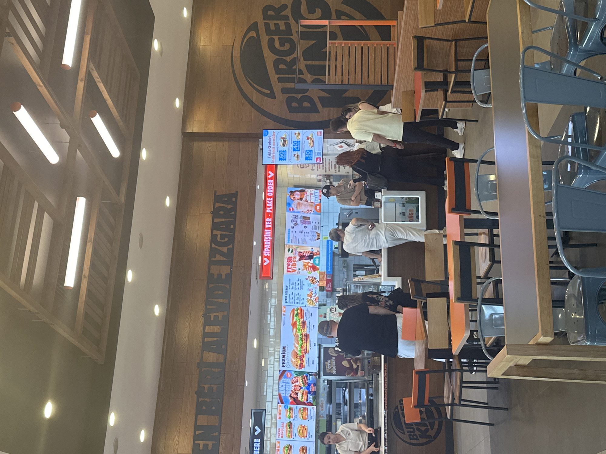 The image shows the interior of a Burger King restaurant, likely located in Manisa, Turkey, as the Turkish language is prominent. A line of people is visible, waiting to place orders at the counter, which features digital menu boards displaying food options. The restaurant design includes wood paneling, Burger King branding, and modern lighting. Seating is available at high tables with blue chairs. People appear to be engaged in the act of ordering food.