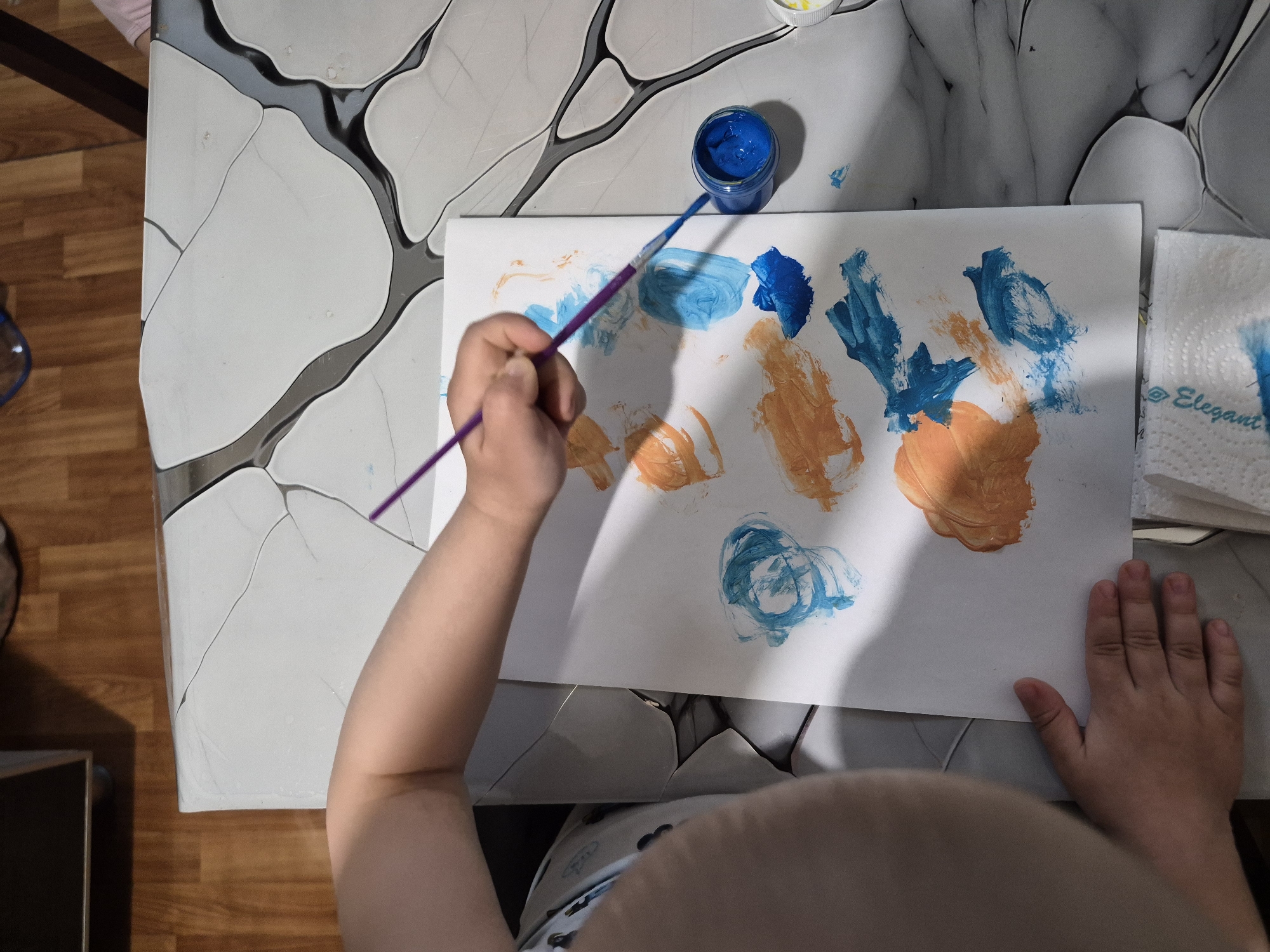 A child is painting on a white sheet of paper. The paper is on a table with a marbled pattern. The child is using a purple paintbrush and blue paint from a small jar. There are orange and blue paint strokes on the paper. A stack of white paper towels labeled "Elegant" is also visible. The child's torso and hands are visible in the frame. The environment appears to be an indoor setting. The city of Tobolsk, Russia, is not discernible from the image.