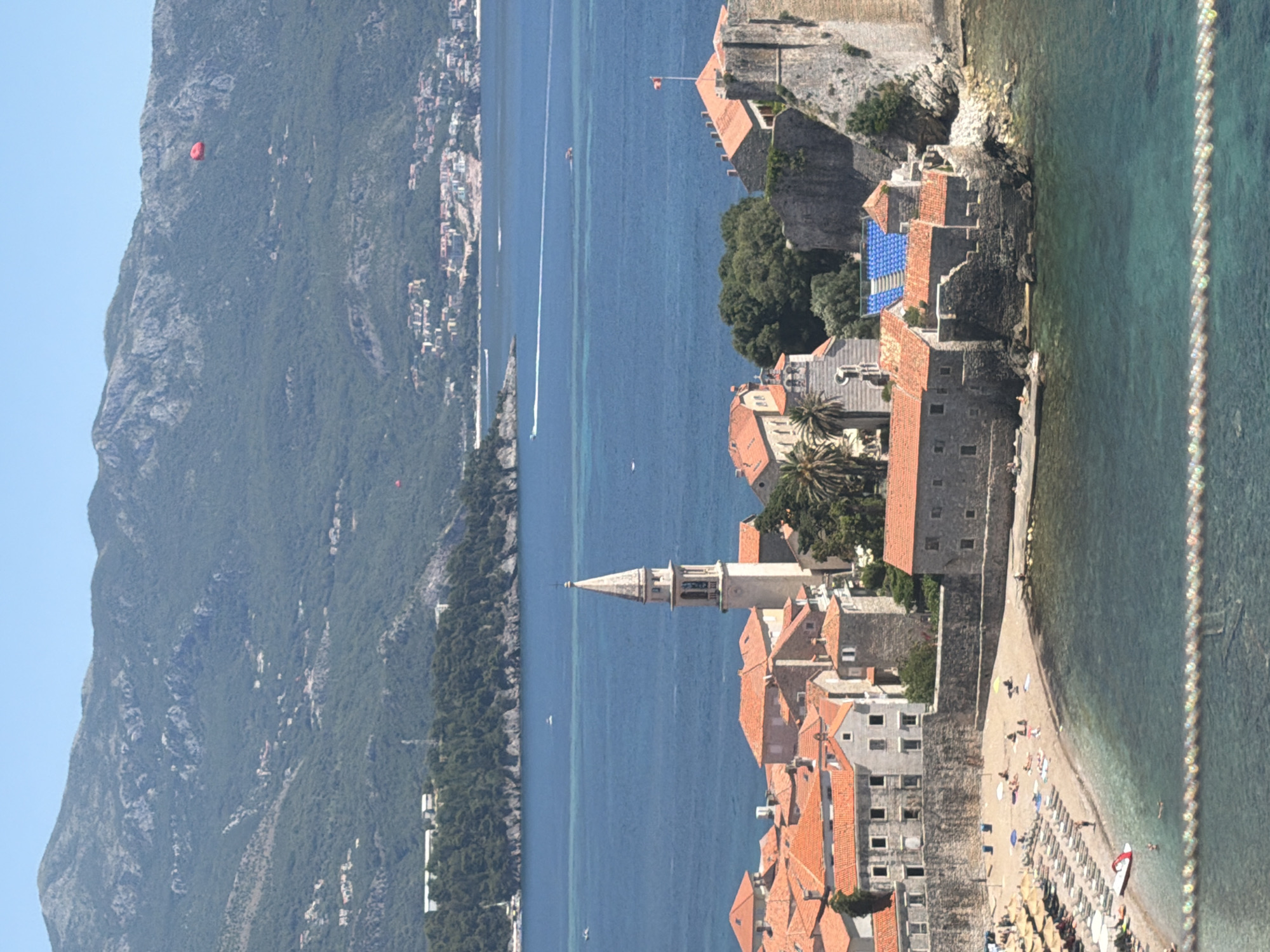 This image provides an aerial view of the historic coastal city of Budva, Montenegro. The scene is dominated by a clear, light blue sky above a range of substantial, green-forested mountains with visible rocky outcrops.
The Adriatic Sea occupies the foreground and middle ground, displaying a gradient from deep blue in deeper areas to clear turquoise near the shore. Multiple white wakes on the water indicate the presence and movement of small boats or personal watercraft. High in the sky, over the sea and in front of the mountains, a single red parasail is prominently visible.
The focal point is the Old Town of Budva, situated on a small peninsula. It features a dense cluster of buildings characterized by reddish-orange tiled roofs and light-colored stone or stucco facades. A tall, light-colored church steeple with a conical spire rises centrally within the old town's architecture. Portions of fortified stone walls are visible along the seaward edges, particularly on the right side of the old town structure. Within these walls, an outdoor venue with visible blue stadium-style seating is present.
To the left of the old town, a crescent-shaped sandy beach is visible. This beach is equipped with numerous rows of straw umbrellas and sun loungers. Approximately two dozen individuals are discernible on the beach and in the adjacent water, engaged in activities such as sunbathing, walking, and swimming. A red inflatable watercraft rests near the water's edge on the beach.
Beyond the old town and along the coastline in the background, residential buildings with red roofs are scattered across the lower slopes of the green mountains.