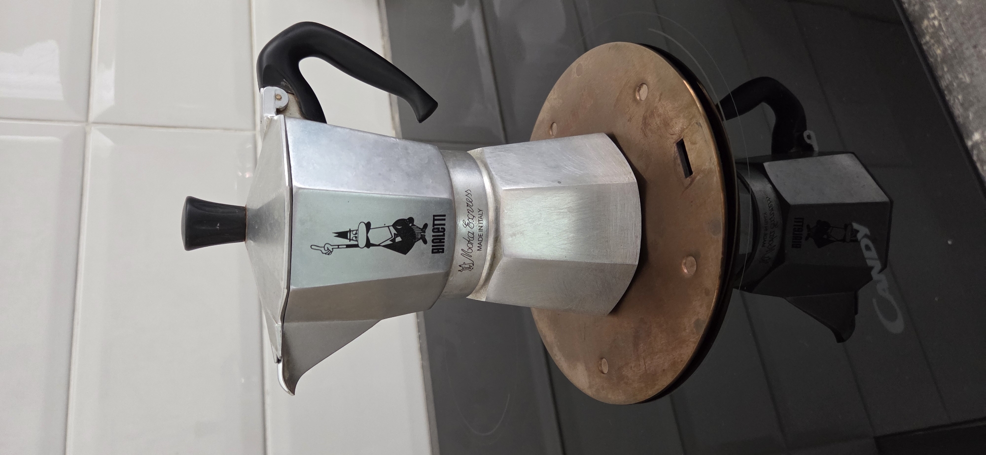 A Bialetti Moka Express stovetop espresso maker is positioned on a copper heat diffuser placed on a black glass electric stovetop. The espresso maker is made of aluminum and features the iconic Bialetti man logo and "Moka Express MADE IN ITALY" text. The stovetop has a white tiled backsplash behind it. The espresso maker is reflecting in the glass surface of the stovetop, showing the "Candy" brand logo. The scene is indoors, likely in a kitchen.