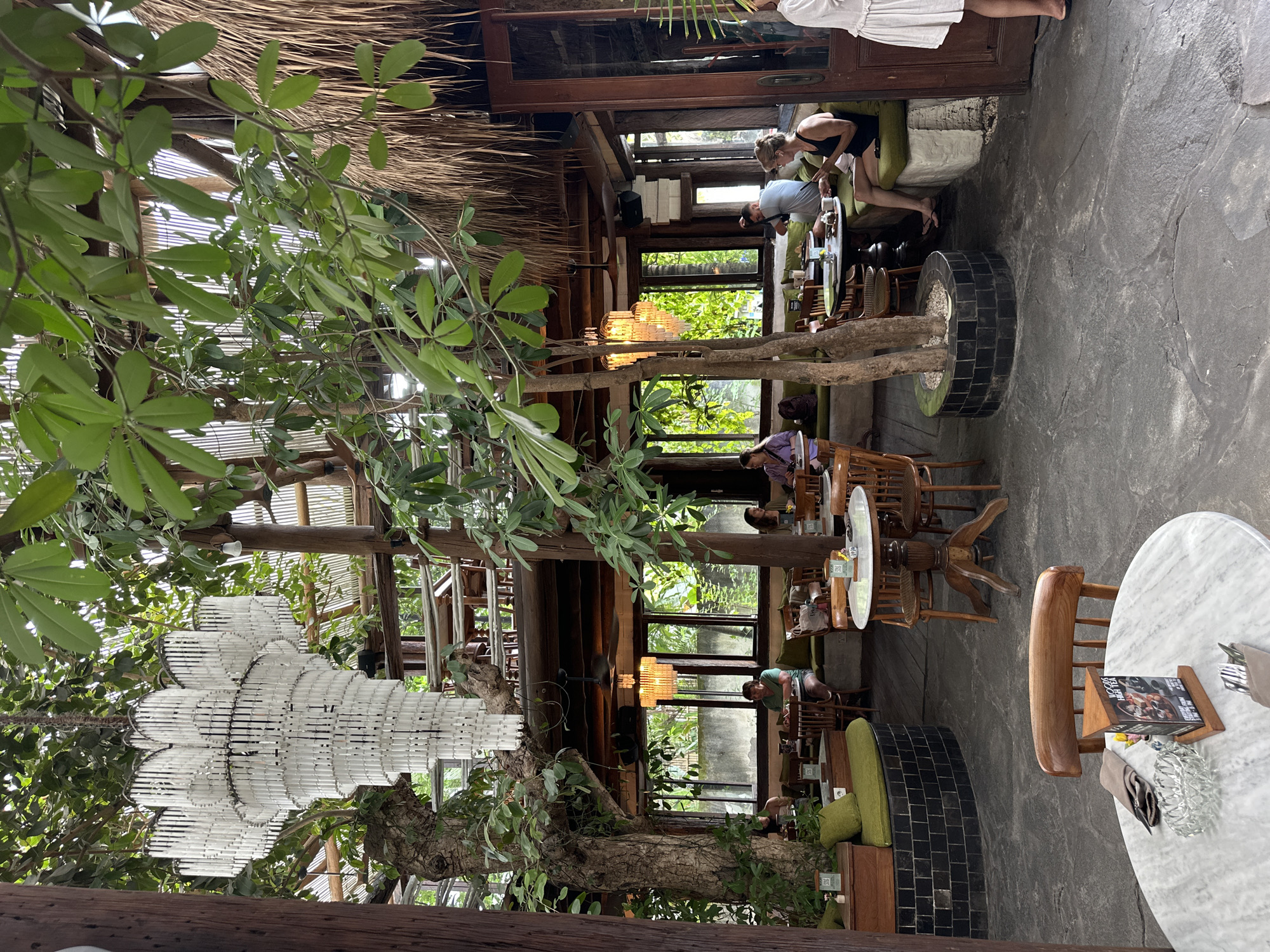This indoor shot of a dining area features multiple tables with wooden chairs, trees growing in the center of the room, and a large chandelier. Patrons sit and eat at the tables. Outside, the windows show green trees and gray buildings. The location is identified as Mengwi, Indonesia.