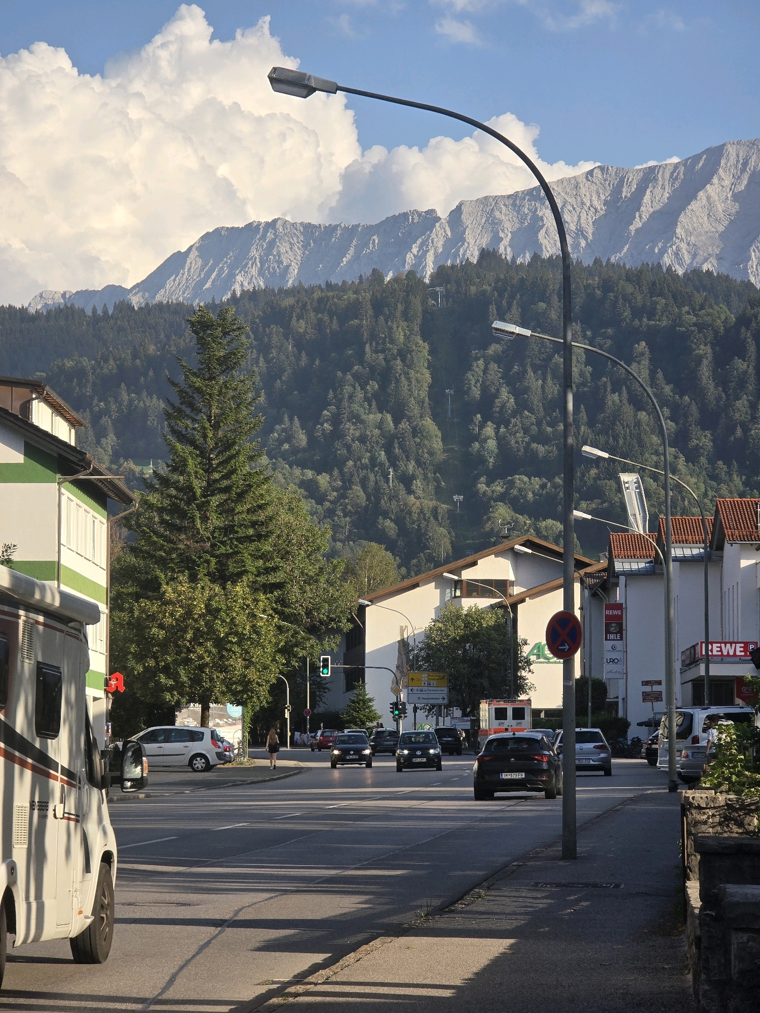 The image depicts a street scene in Garmisch-Partenkirchen, Germany, on a sunny day with dramatic clouds and a mountain range in the background. A white camper van is parked on the left side of the road. Several cars are driving on the multi-lane asphalt road, which is bordered by sidewalks and buildings. Traffic lights, streetlights, and road signs are visible. The buildings on the right side of the street include commercial establishments, one of which has a "REWE" sign. A person is walking on the sidewalk. The background features a densely forested mountain slope, with a cable car visible ascending the mountain.