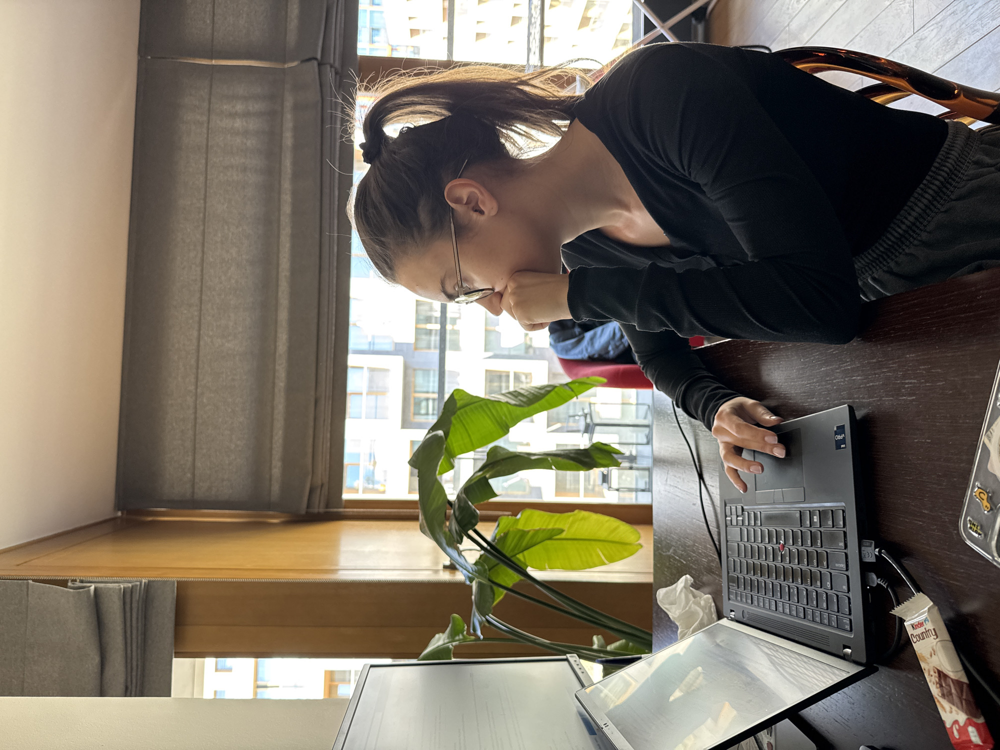 A woman with dark hair tied in a high ponytail and wearing glasses is seated at a dark wooden desk, engaged in computer work. She is viewed from the side, leaning forward with her right hand on a laptop's touchpad and her left hand touching her chin, suggesting concentration or contemplation. She wears a black long-sleeved top and dark sweatpants.
On the desk in front of her is an open black laptop, which has a "VPRO" sticker visible. A USB-C hub is plugged into the laptop. To the right of the laptop, a Kinder Country chocolate bar rests on the desk, alongside a crumpled tissue. Another monitor or open laptop is partially visible in the foreground, indicating a dual-screen setup.
Behind the woman, a large window provides ample natural light. Grey blinds or curtains are partially drawn, but the window offers a view of modern, multi-story urban buildings, typical of an apartment complex in a city like Warsaw, Poland. A large houseplant with broad, vibrant green leaves (resembling a Strelitzia or Bird of Paradise plant) stands between the desk and the window. The bright sunlight suggests it is daytime with clear weather. The back of a chair with an ornate, possibly tortoiseshell-patterned frame is visible behind her.