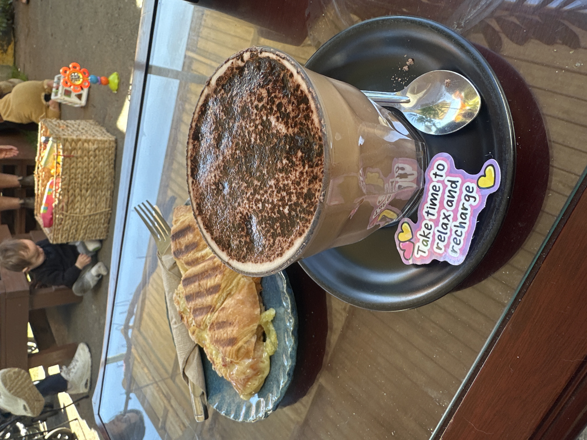 A close-up, eye-level shot shows a coffee cup with a spoon on a saucer. The cup is filled with a chocolate-colored beverage dusted with cocoa powder. A sticker with the text "Take time to relax and recharge" adorns the saucer. Next to the cup is a plate with a grilled pastry. In the background, a child sits on the ground next to a woven basket, and a person's legs are visible, wearing white sneakers and dark pants. The setting appears to be an outdoor cafe, possibly in Bellingen, Australia, with a wooden table and a glass surface.