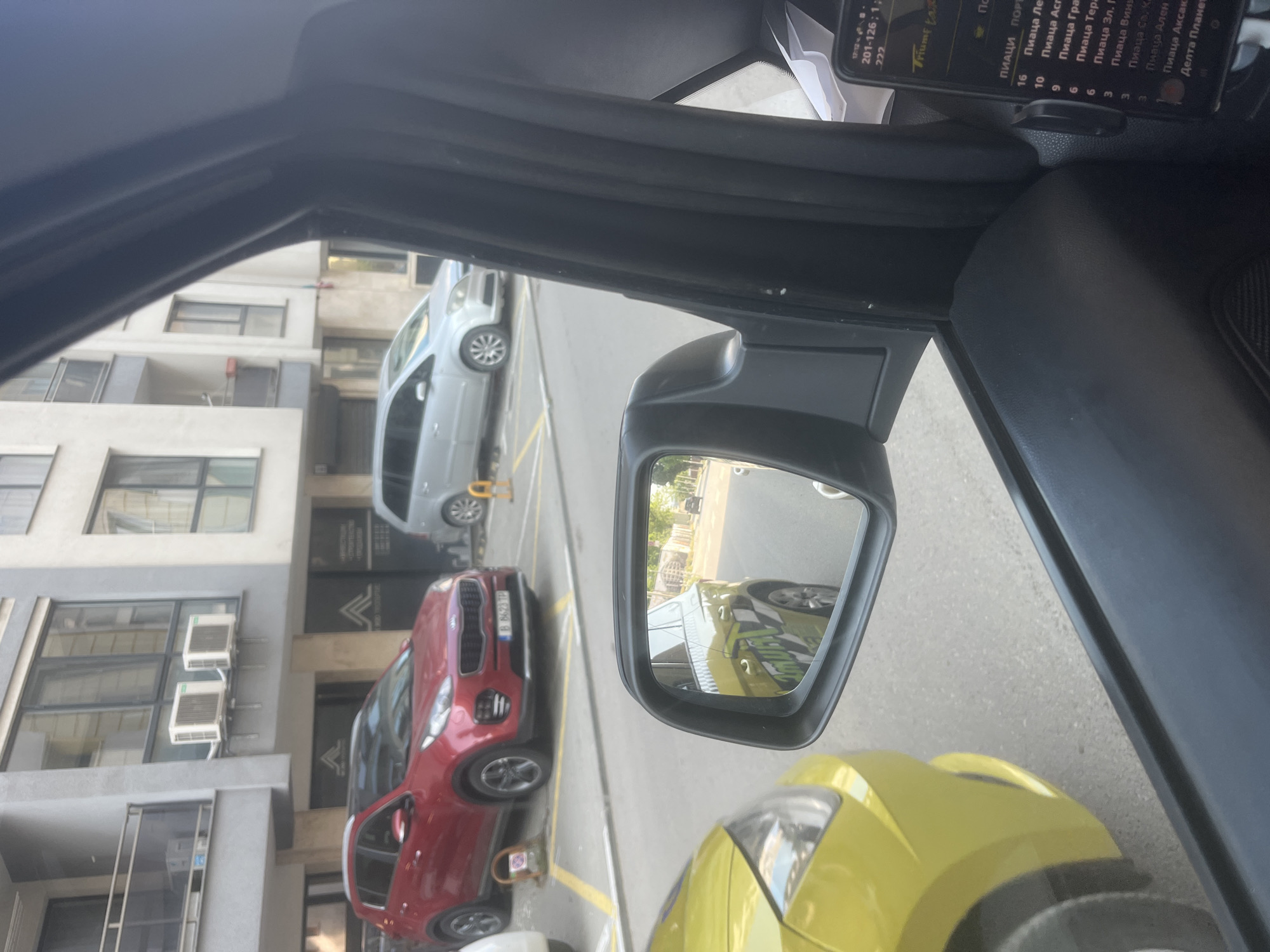 The image, taken from inside a vehicle, captures a daytime urban street scene in Varna, Bulgaria. The perspective is from the front passenger side, looking out towards buildings and parked cars.
In the immediate foreground, part of a yellow car is visible on the right, likely the vehicle from which the photograph was taken. The car's side mirror is prominent, reflecting a yellow vehicle with "Triumf" branding, suggesting a taxi service.
Beyond the vehicle, a red Kia Sportage SUV (license plate B 8628 TP) is parked on the street, followed by a light gray car further down. The street features yellow parking lines. On the left, multi-story buildings with numerous windows and balconies line the road. Several external air conditioning units are mounted on one of the building's facades.
Inside the car, a smartphone is mounted on the dashboard, displaying what appears to be a taxi dispatch or navigation interface. The screen prominently shows "Triumf tax" at the top, along with a list of "ПИАЦИ" (Piazi - likely 'Squares' or 'Locations' in Bulgarian), including "Пиаца Ле" (Piaza Le), "Пиаца Асг" (Piaza Asg), "Пиаца Гра" (Piaza Gra), "Пиаца Тера" (Piaza Tera), "Пиаца Зл. Г" (Piaza Zl. G), "Пиаца Вини" (Piaza Vini), "Пиаца Св. Ка" (Piaza Sv. Ka), "Пиаца Ален" (Piaza Alen), "Пиаца Аксак" (Piaza Aksak), and "Делта Планет" (Delta Planet), each paired with numbers (e.g., 16, 10, 9, 6). Additional visible text on the screen includes "201-126;1;" and "222".
The scene depicts a common urban environment, likely from a parked taxi, with clear weather conditions.