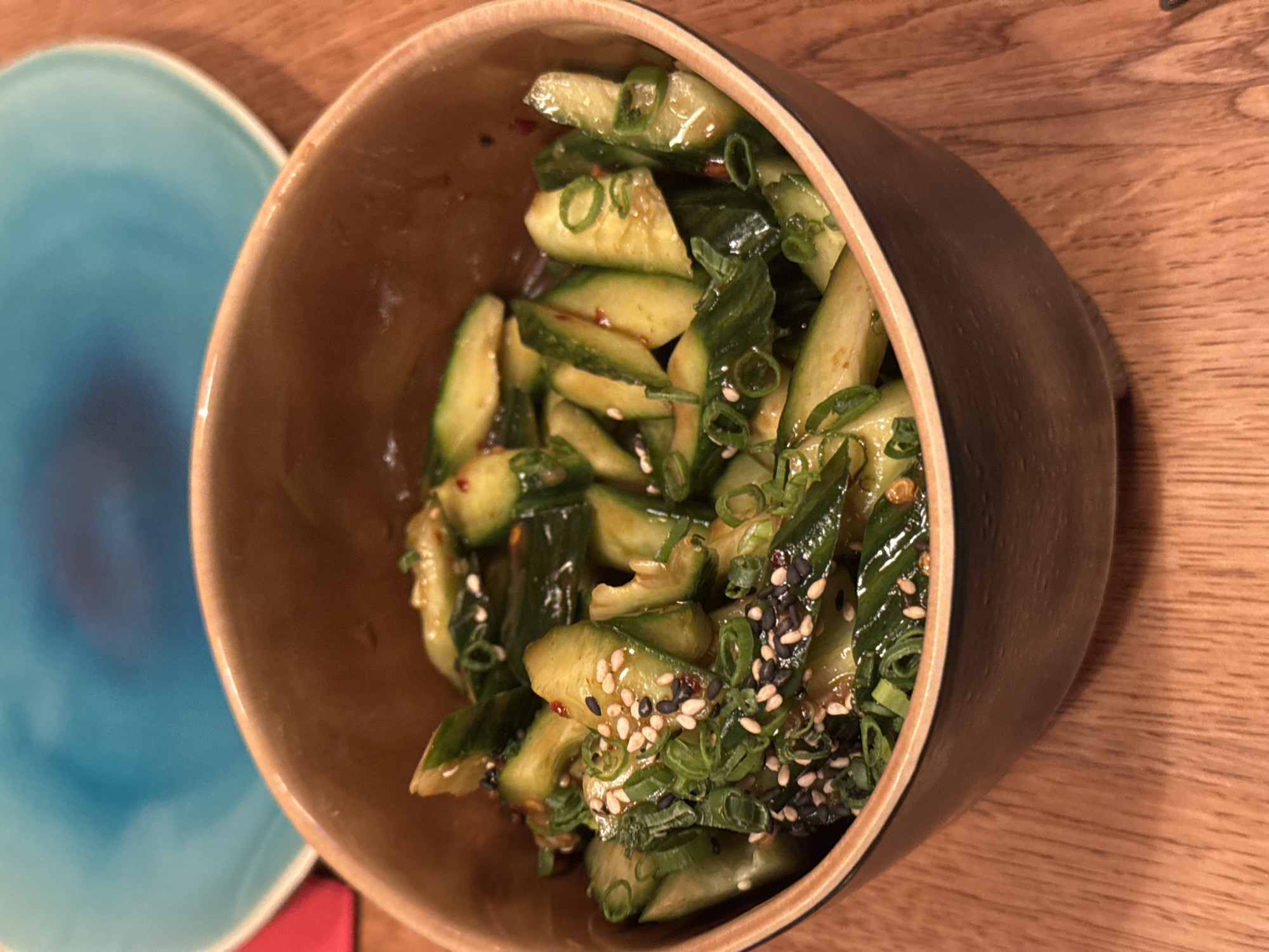 A medium shot depicts a bowl of cucumber salad, likely prepared in an Asian style. The cucumber pieces, cut into angled shapes, are mixed with sesame seeds (both black and white) and sliced green onions, coated in a glaze. The bowl is made of wood, with a light-colored interior and a dark brown exterior. Adjacent to the bowl is a light blue plate. The background consists of a wooden table. The city and country of the scene (Sofia, Bulgaria) are not visible in the image.