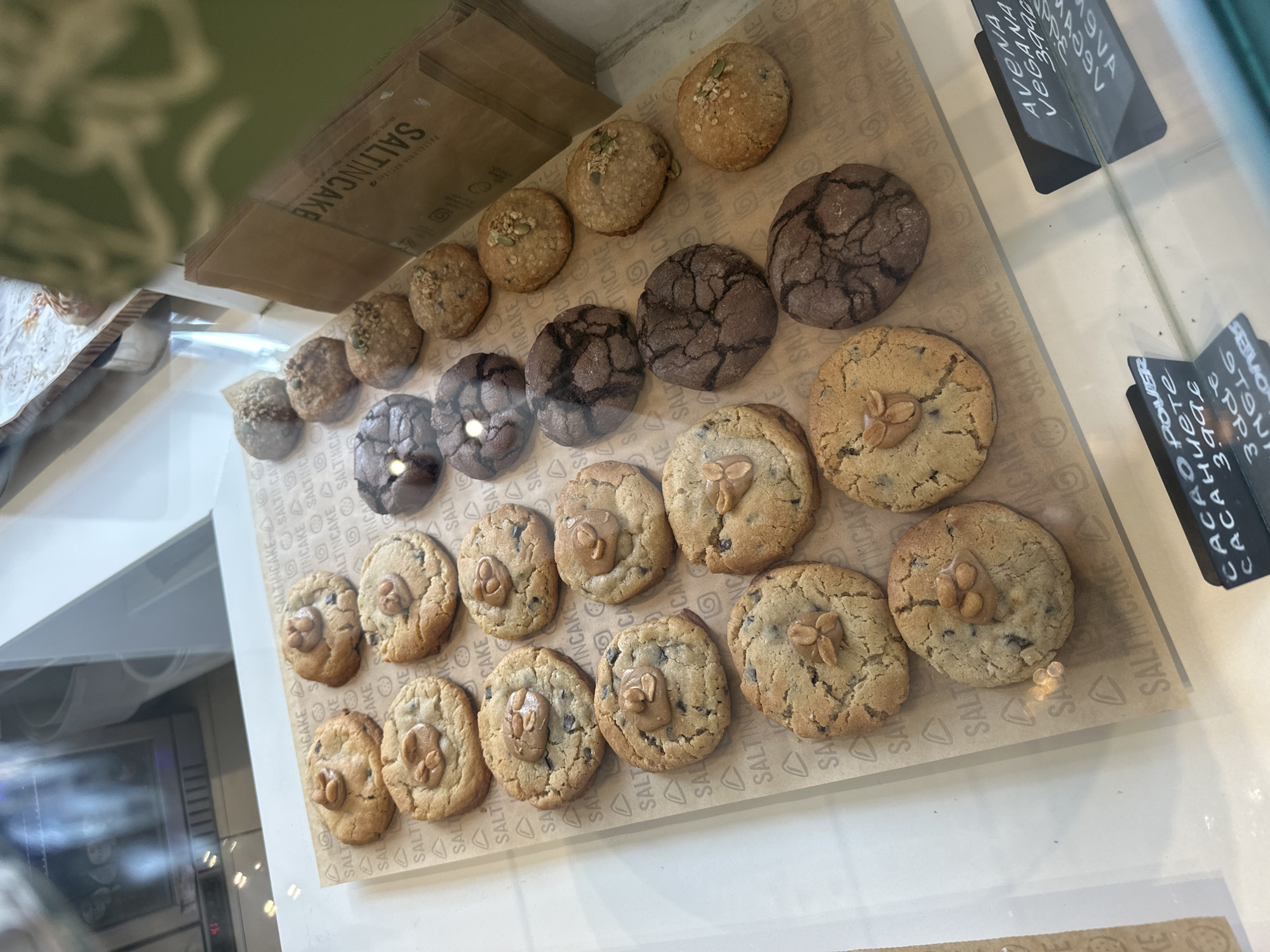 The image displays a variety of cookies presented in a glass display case, likely within a bakery or cafe. The cookies are arranged in rows on patterned brown paper liners, which are imprinted with the brand name "SALTINCAKE" multiple times. In the background, stacked brown paper bags also feature the "SALTINCAKE" logo and the text "PASTELERÍA NATURAL" (Natural Pastry Shop).
Several types of cookies are visible. Many are light-colored, possibly chocolate chip, and are topped with a distinct dollop or cluster of what appears to be peanut butter or nuts. There are also dark brown cookies with a characteristic crinkled surface, resembling chocolate crinkle cookies. Further back, another variety of lighter cookies is topped with seeds, such as pumpkin or sunflower seeds.
Two black information tags are visible. One clearly states "AVENA VEGANA" (Oat Vegan) along with the numbers "3996" and "4764". The second tag reads "CACAO POWER" followed by "CACAHUETE" (Peanut), "3996", "3dde", and other fragmented text including "ここの上の" and "MERS". Reflections on the glass display case cause some minor visual obscuration. The scene suggests an inviting presentation of freshly baked goods emphasizing natural ingredients.