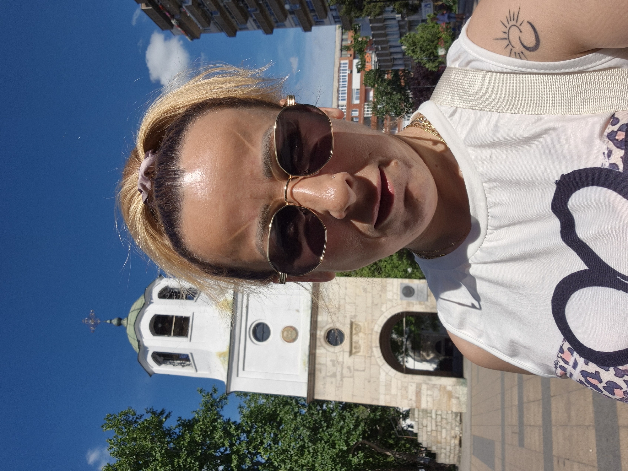 A medium shot captures a woman taking a selfie outdoors on a sunny day. She has blonde hair pulled back in a ponytail and is wearing sunglasses and a white sleeveless top. Her left arm, visible on the right side of the frame, bears a tattoo of a crescent moon and a sun with rays.
In the background, a white clock tower with a green dome stands against a clear blue sky with a few white clouds. The tower has arched openings and a cross at the apex. To the left of the tower is a lush green tree. Further in the background, to the right of the tower, are multi-story buildings. The ground is paved with square tiles. The woman's facial expression is neutral.