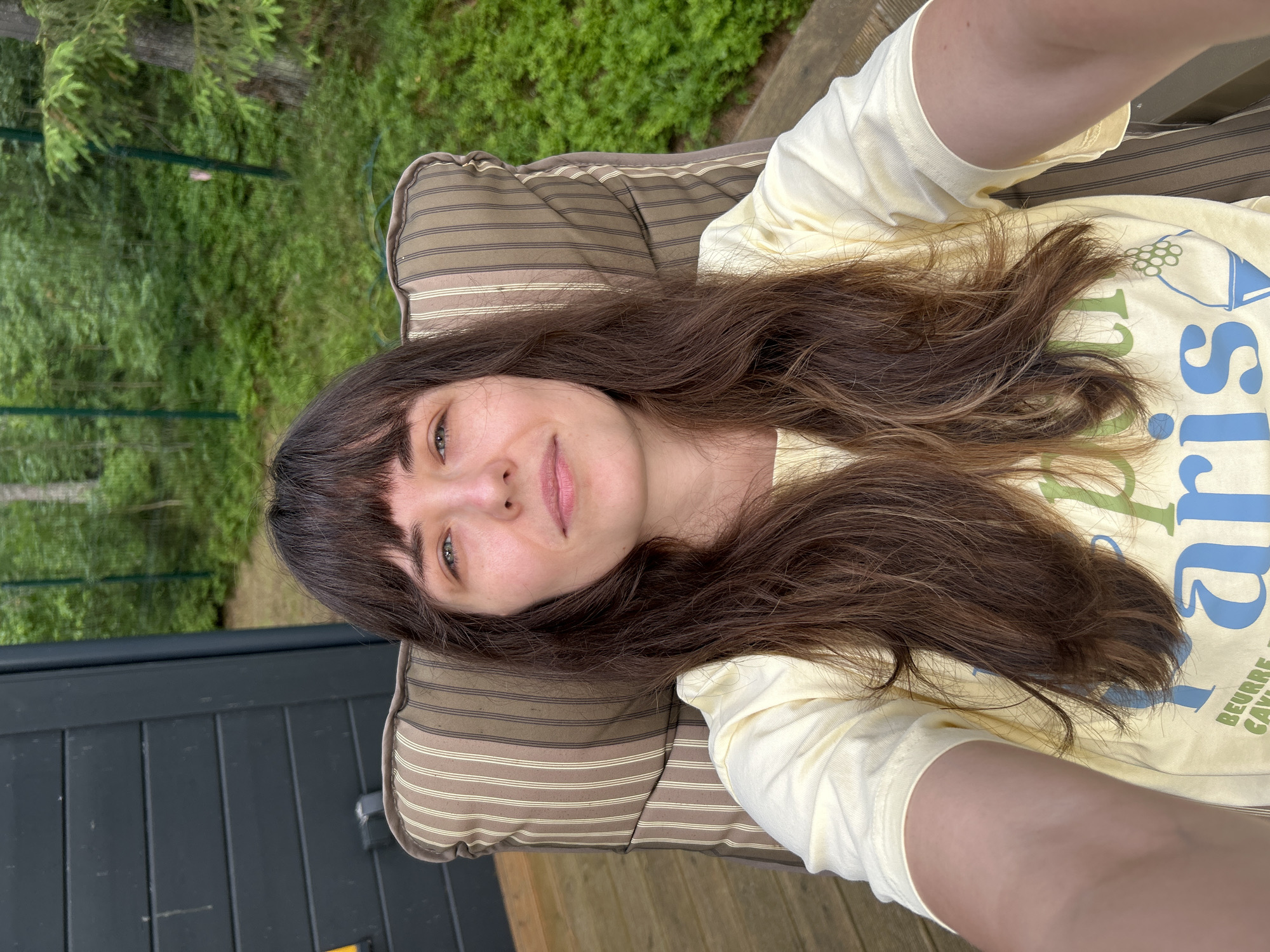 A woman with long brown hair and bangs sits in a striped outdoor chair on a wooden deck. She wears a pale yellow t-shirt with blue and green lettering. Behind her, a dark wooden wall of a building is visible on the left, and lush green foliage, including trees and bushes, fills the background. A green fence is also present in the background. The image appears to be a selfie taken outdoors. The location is stated to be Shepelyovo, Russia.