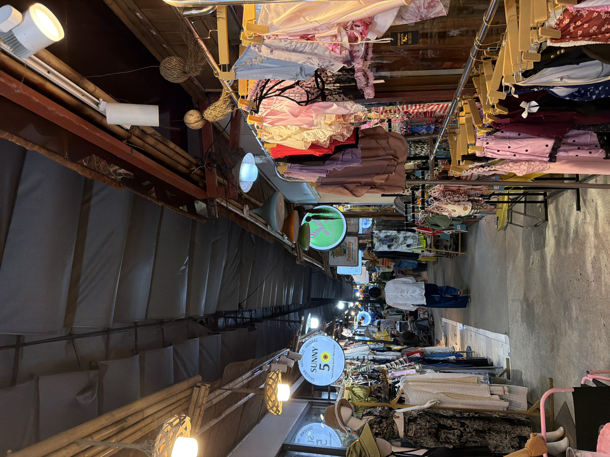 The image depicts a narrow market alleyway in Đà Nẵng, Vietnam. Racks of clothing are densely arranged on either side, with various garments on hangers and shelves, including lingerie, tops, and dresses. Signs are visible, including one that says "Sunny 5: Spreading good vibes only" and other brands like "PalmBoy" and "Midnight Secrets". Several people are seen walking through the alleyway. The setting has a cluttered yet organized feel, typical of street markets in Southeast Asia. The alleyway is covered, and there are lights overhead.