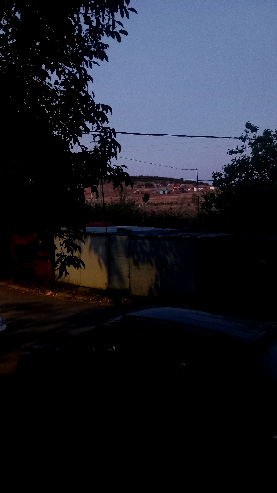 A dimly lit exterior scene captures the twilight sky above a residential hillside dotted with small buildings. In the foreground, dark silhouettes of trees frame the view. A concrete wall, partially illuminated by an unseen light source, runs horizontally across the midground, casting shadows. A portion of a dark-colored vehicle is visible in the lower right corner, its shape indistinct due to the low light. The overall atmosphere is quiet and subdued, indicative of early evening.